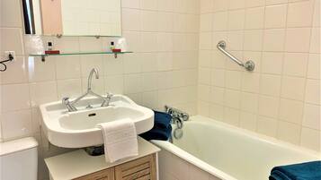 A bathroom with a white sink, a mirror, a glass shelf with toiletries, a bathtub, and a wooden cabinet.