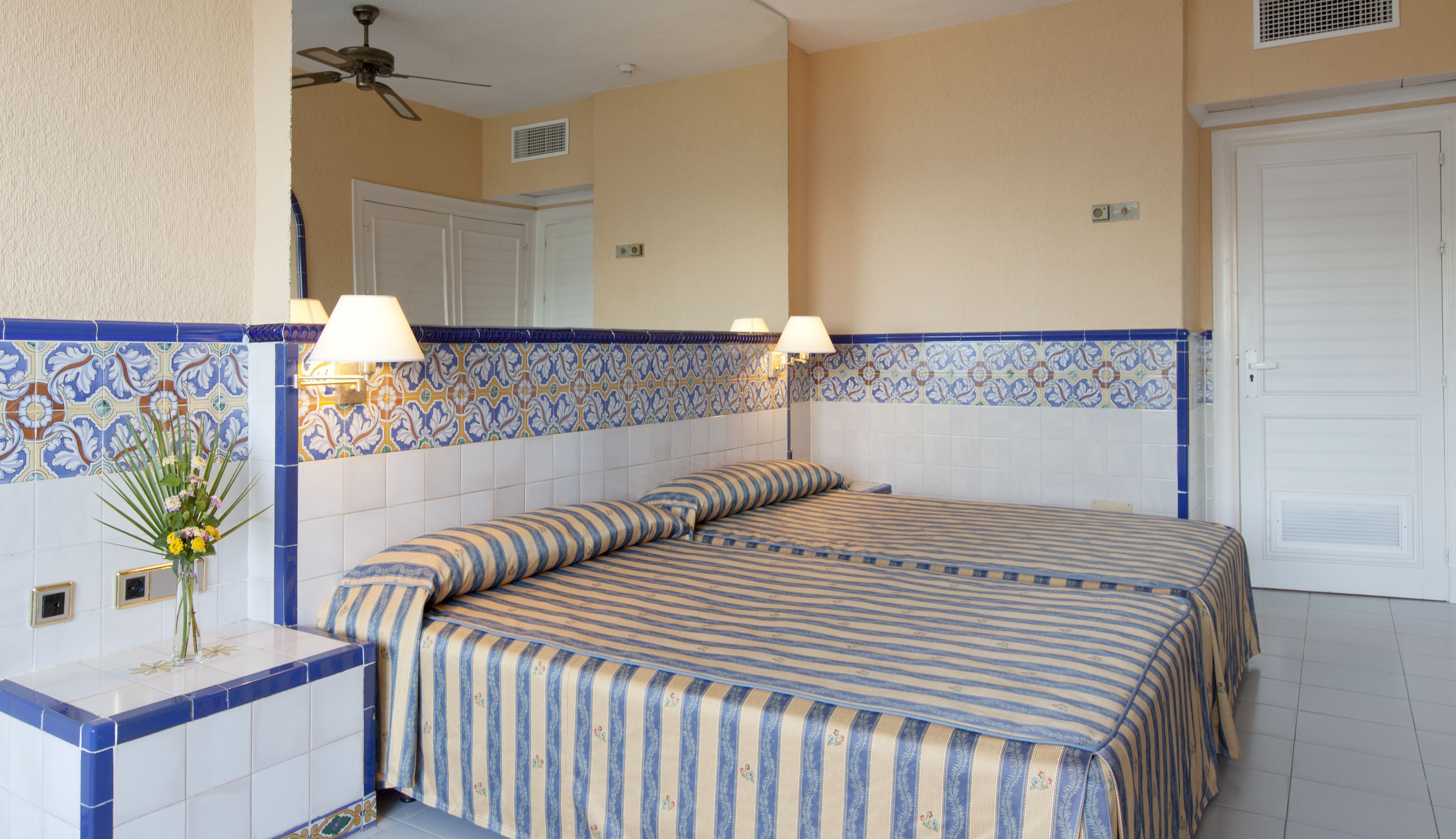 Habitación de hotel con cama, dos lámparas de noche, un espejo y pared de azulejos con diseño azul y blanco.