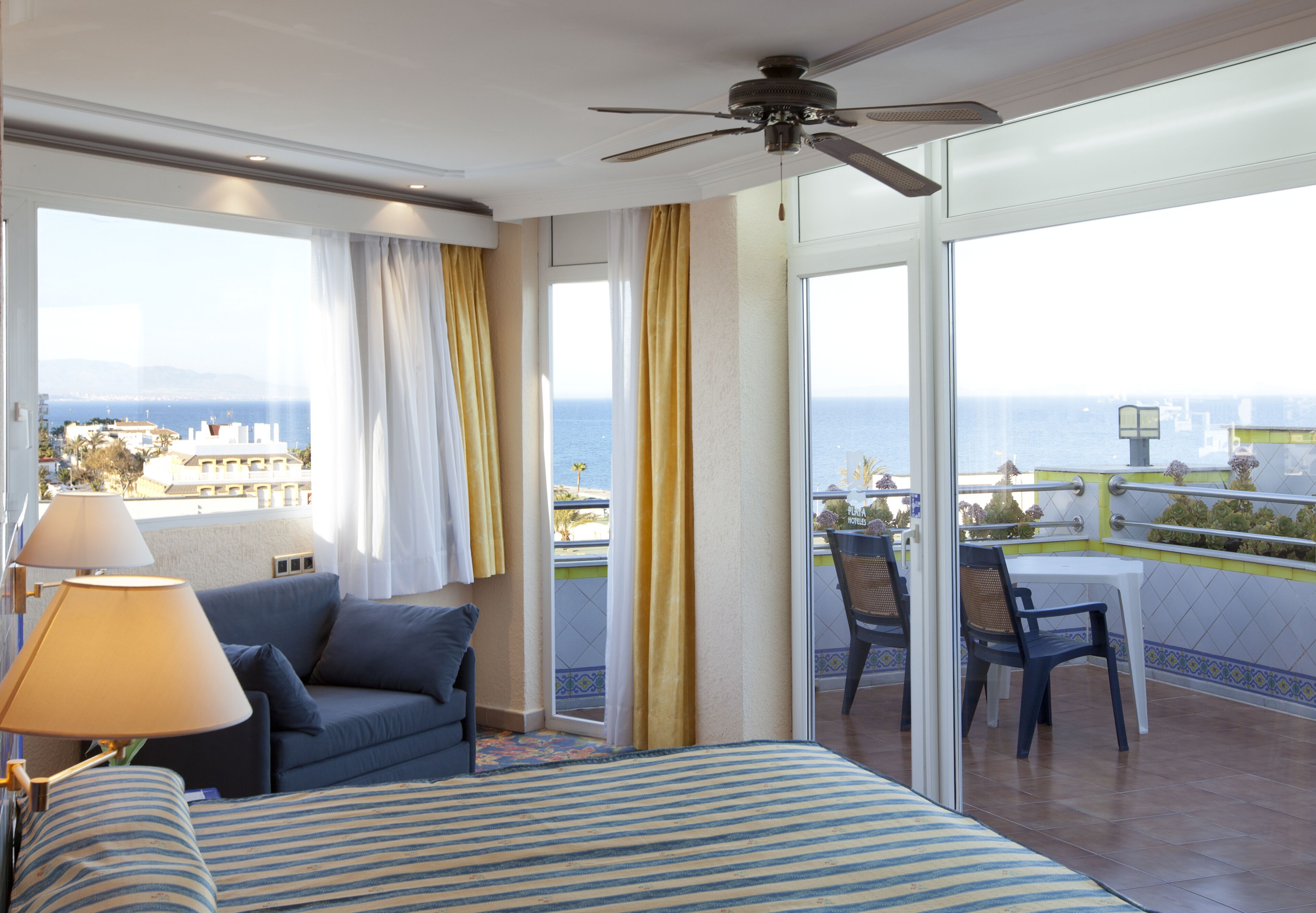 A hotel room with a bed, a sofa, a balcony with a table and chairs, and a view of the sea.