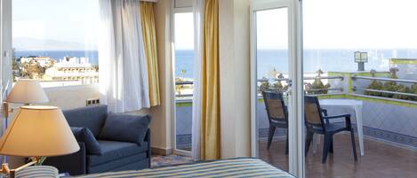 A hotel room with a bed, a sofa, a balcony with a table and chairs, and a view of the sea.