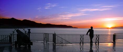 Vista del atardecer desde un paseo marítimo con barandilla de metal y una estatua.