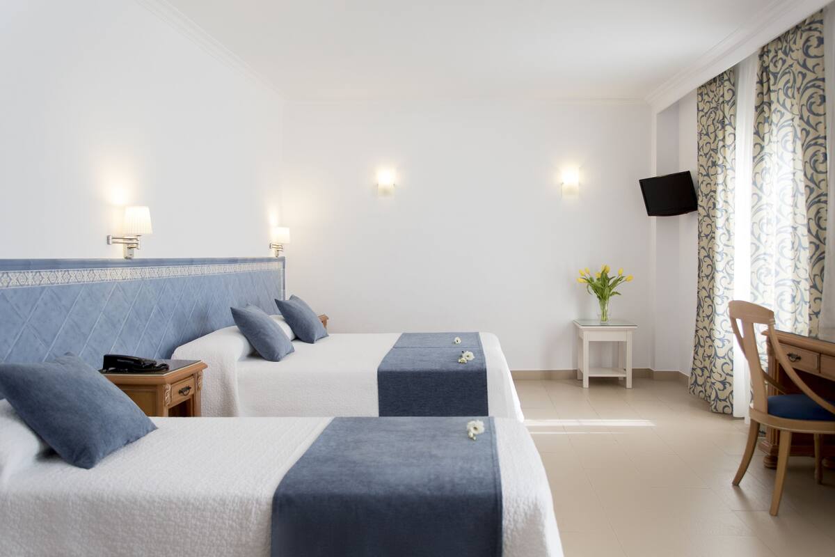 Habitación de hotel con dos camas, un televisor montado en la pared, una mesita con un jarrón de flores y una silla.