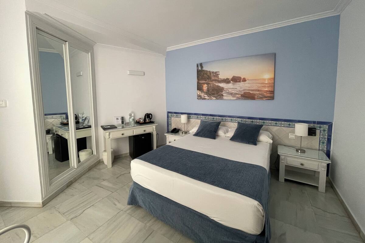 Habitación de hotel con cama, escritorio, silla, espejo, mesita de noche y un cuadro colgado en la pared.