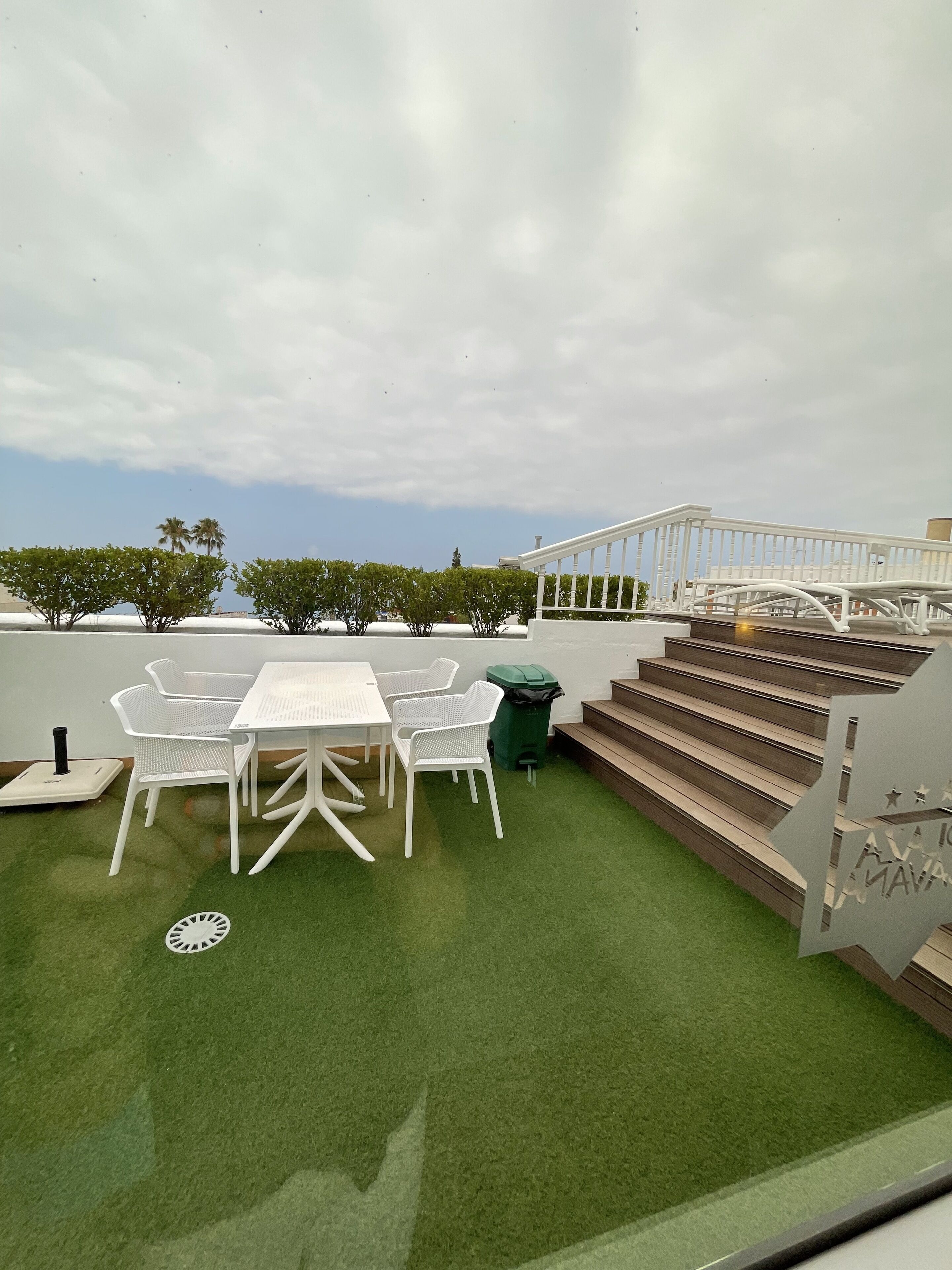 A rooftop patio with a white table and chairs, a green trash bin, and a staircase leading to an upper level.