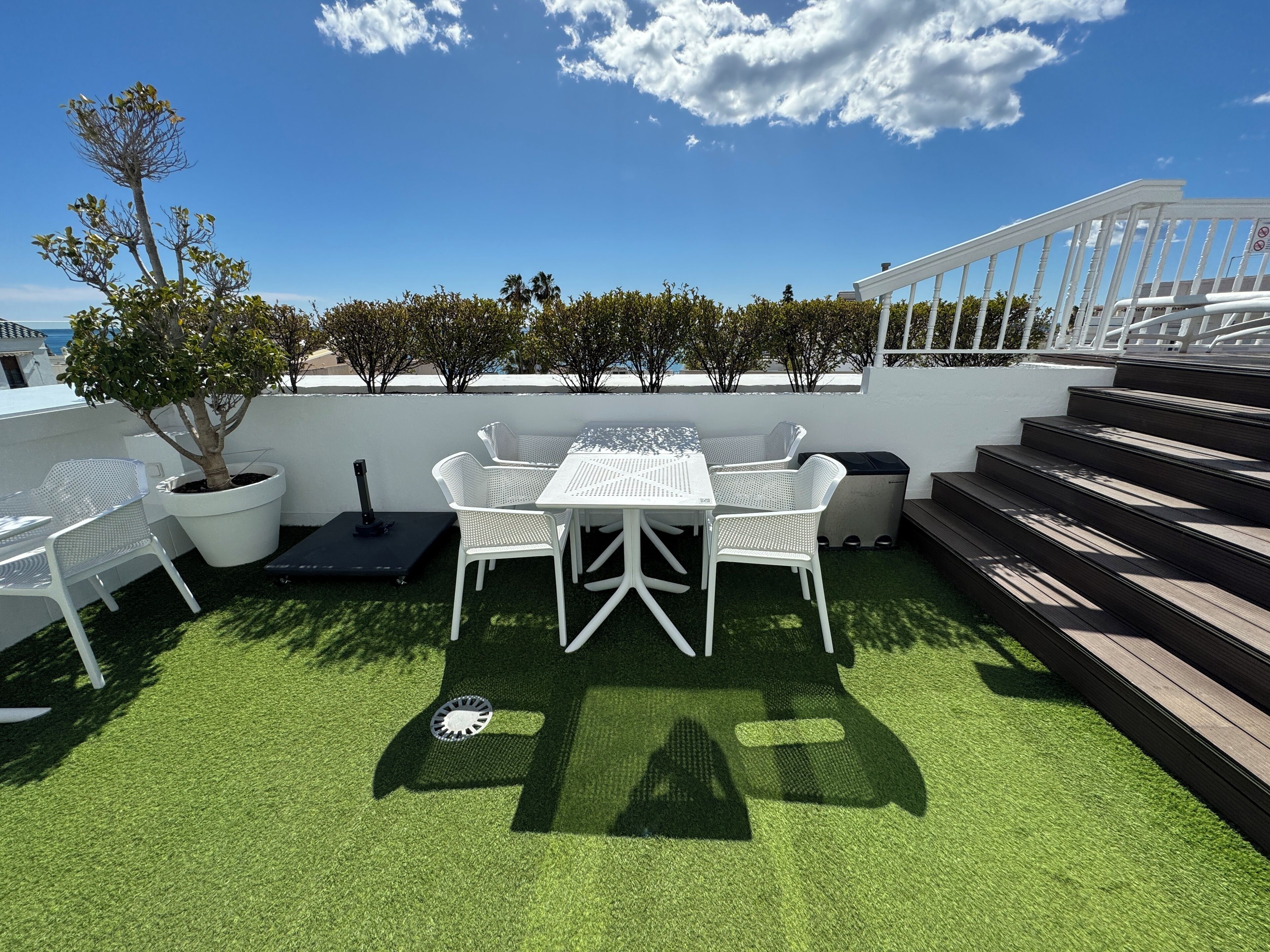 A rooftop patio with a white table and chairs, surrounded by potted plants and a view of the sea.