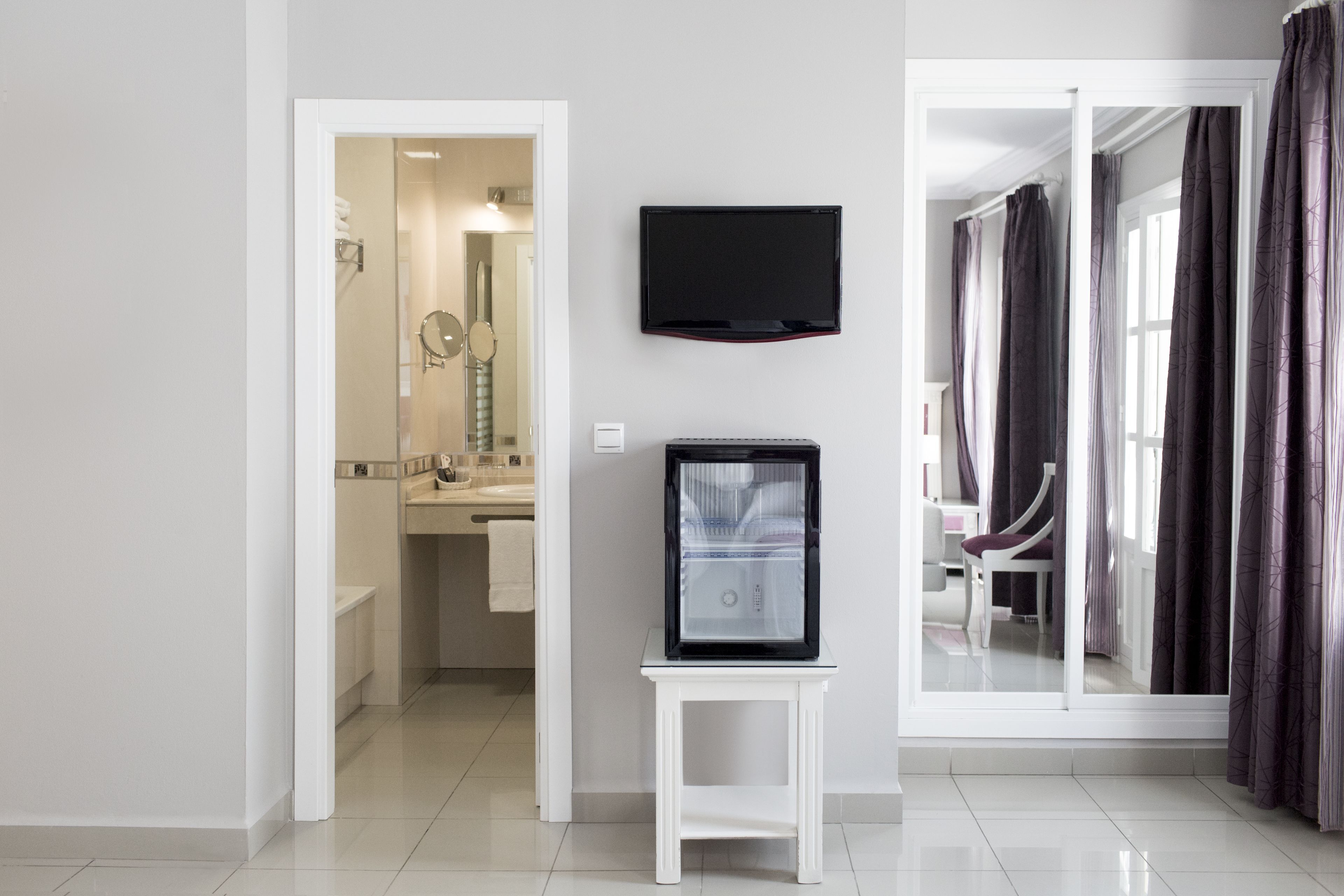 A modern hotel room with a flat-screen TV mounted on the wall, a small table, and a view of the bathroom and balcony.