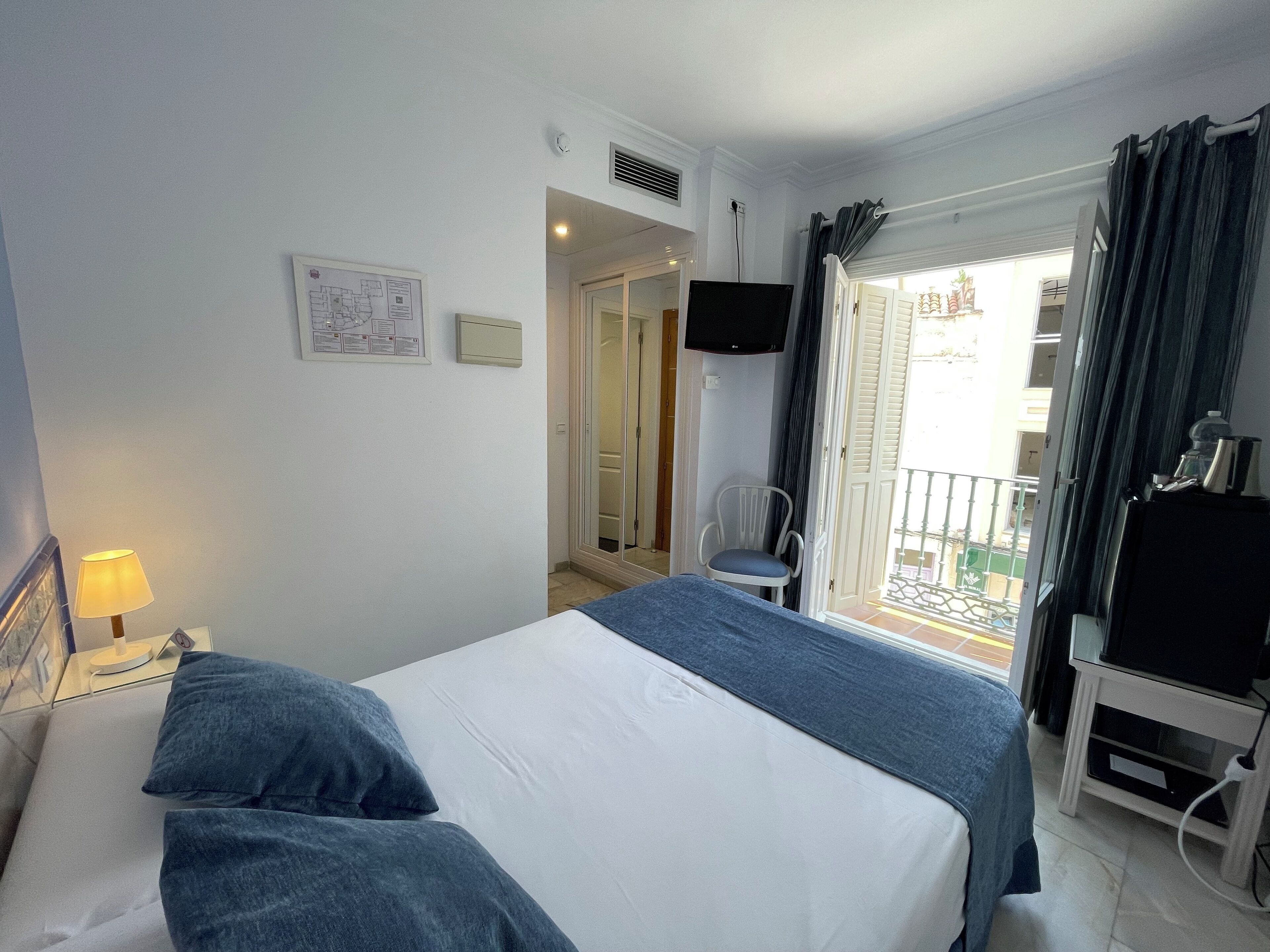 Quarto de hotel com cama, poltrona, televisão e varanda com cortinas.