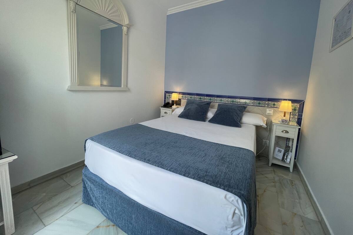 Una cama bien tendida con una colcha azul y blanca, una mesita de noche con una lámpara y un espejo fijado a la pared.
