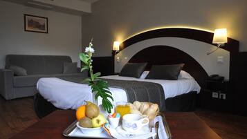Une chambre d’hôtel avec un plateau de petit-déjeuner soigneusement préparé sur la table, un lit avec une tête de lit, une table de chevet avec une lampe, et un canapé gris en arrière-plan.