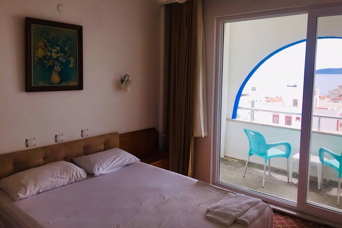 Habitación de hotel con cama, un cuadro, ventana con vistas a edificios y balcón con sillas.