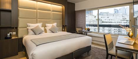 A modern hotel room with a large bed, a desk with a laptop, and a view of the city through large windows.