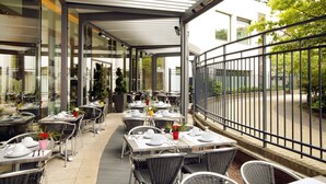An outdoor cafe with tables and chairs, a covered walkway, and a view of the street. - Residhome Courbevoie La Défense (Courbevoie)