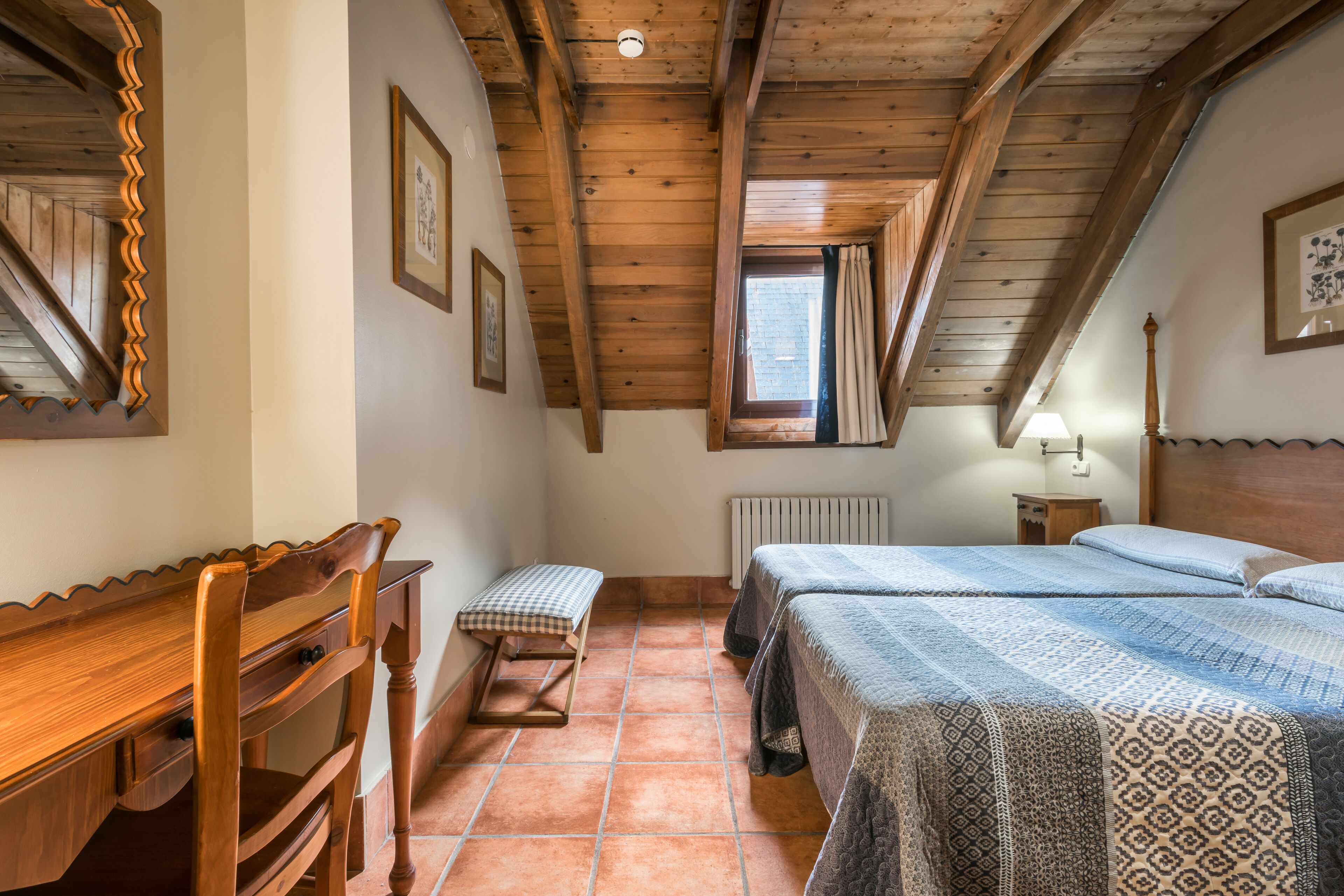 A bedroom with wooden beams, a desk, two beds, a radiator, and a window with curtains.