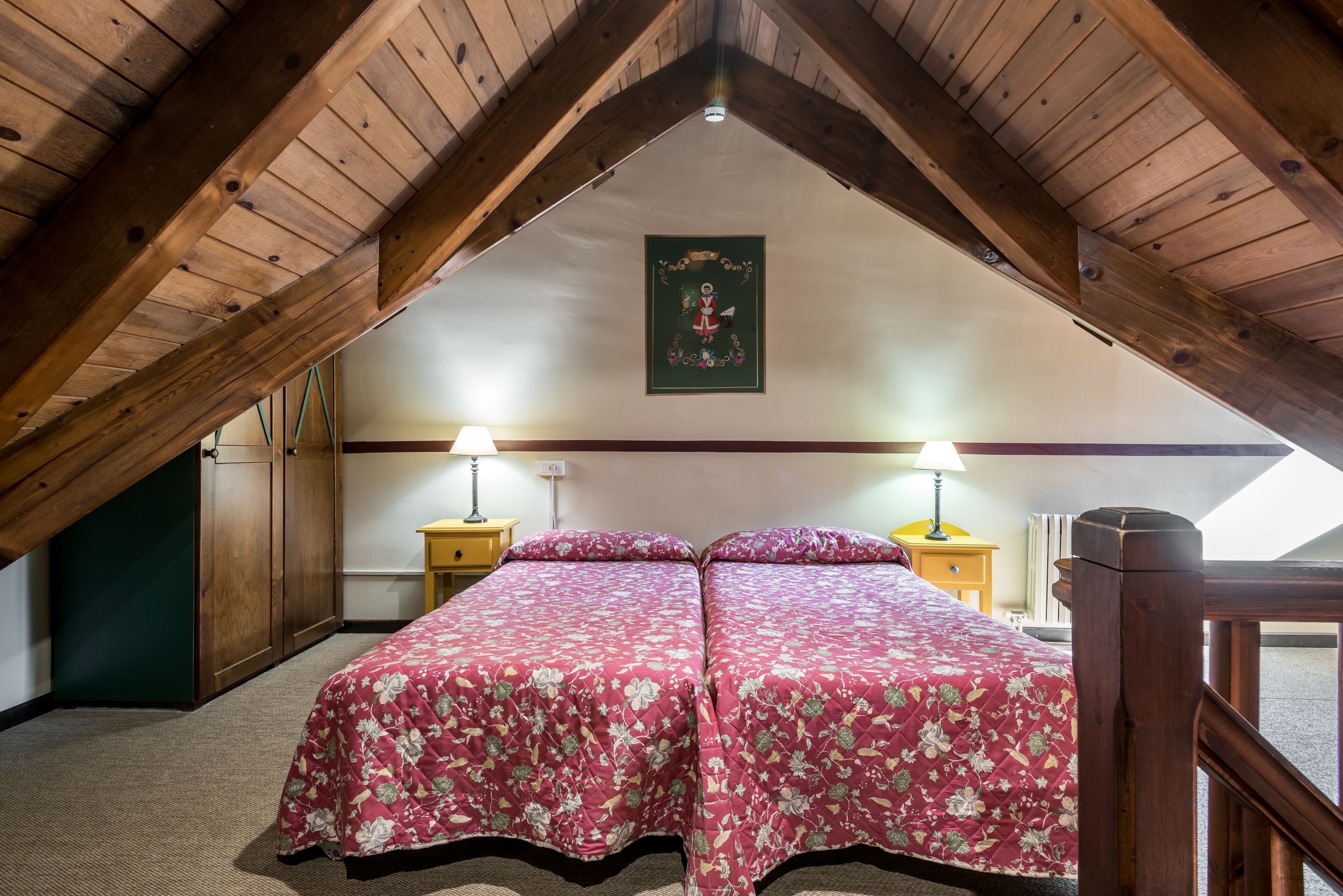 Un dormitorio con techo inclinado, vigas de madera, dos mesitas de noche con lámparas y una cama con ropa de cama floral.