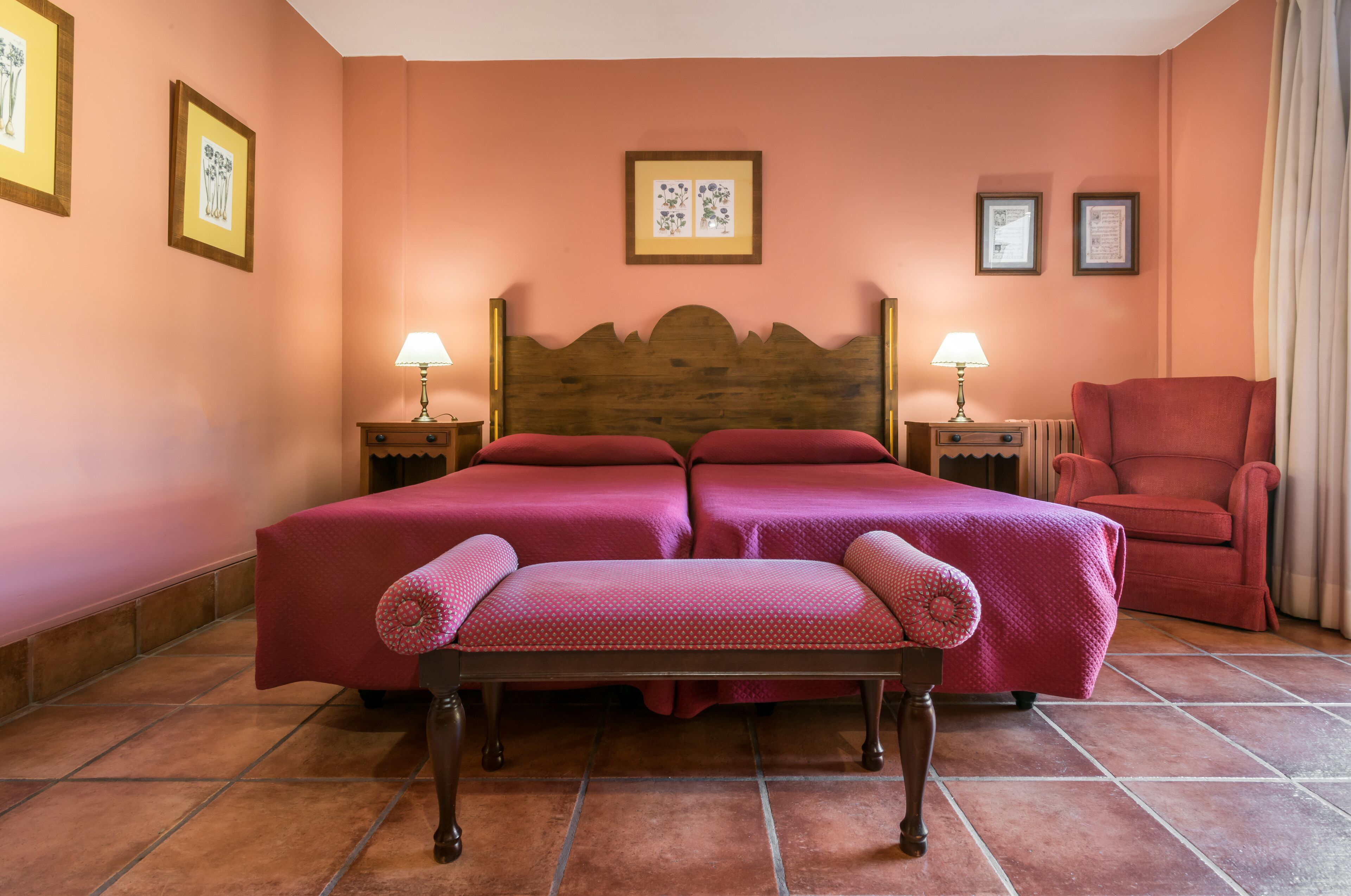 Una cama doble con cabecera de madera, dos mesitas de noche con lámparas, un sillón rojo y un banco.