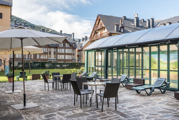 A covered outdoor seating area with tables and chairs, a glass greenhouse structure, and traditional buildings in the background. - El Refugio de Aran Vielha Aparthotel (Vielha e Mijaran)