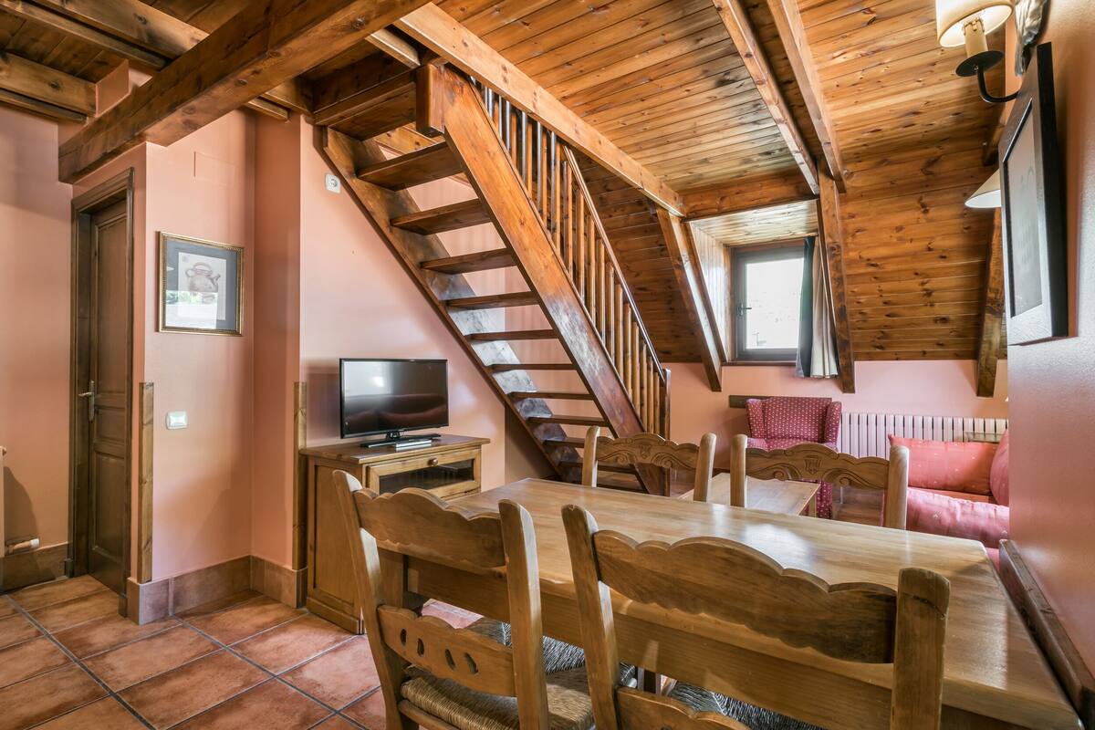 A dining area with a wooden table and chairs, a television on a stand, and a wooden staircase leading to an upper level.