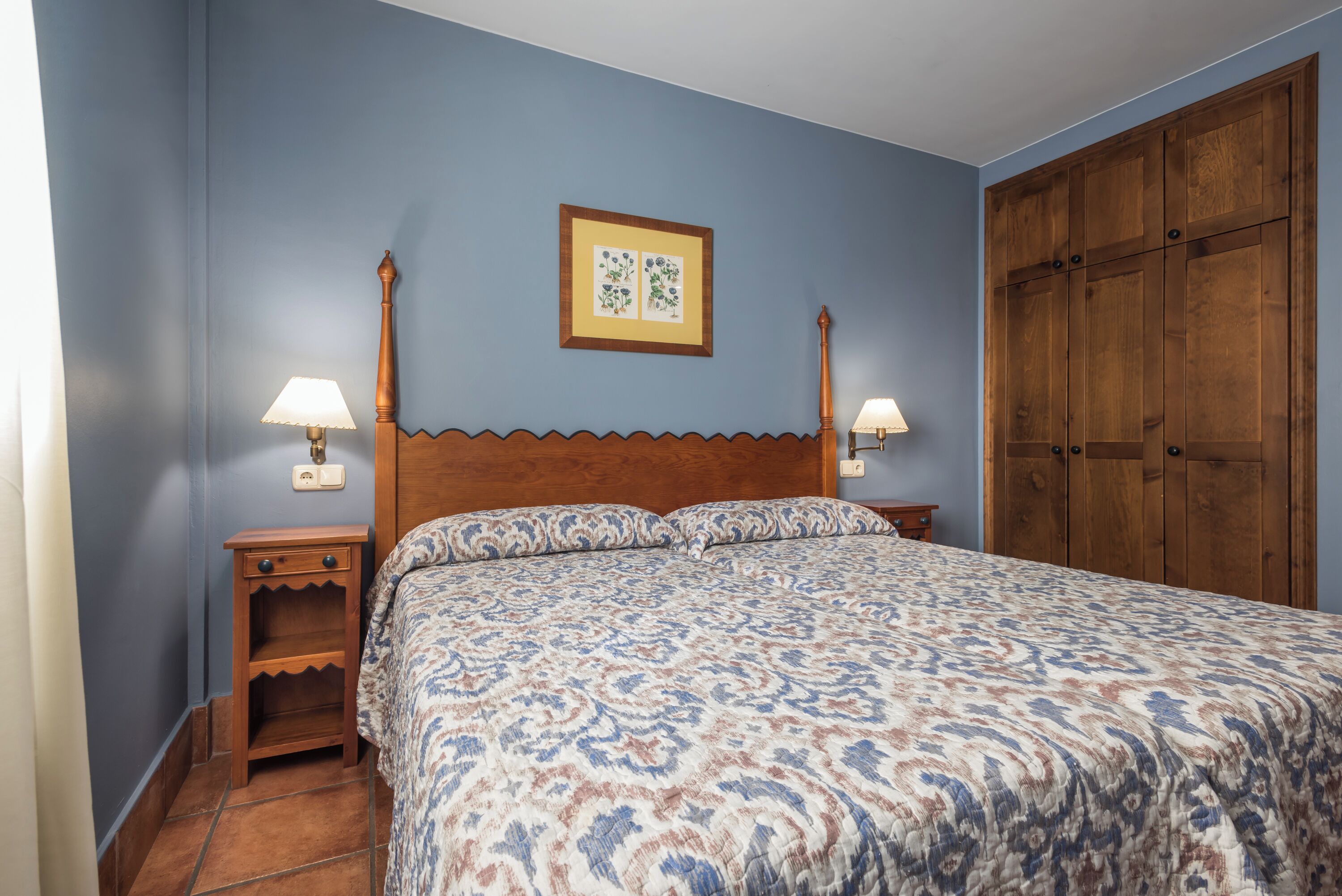 Ein Schlafzimmer mit einem Holzkopfteil, einer gemusterten Bettdecke, zwei Nachttischlampen, einem kleinen Holfnachtkästchen und einem großen Holzschrank.