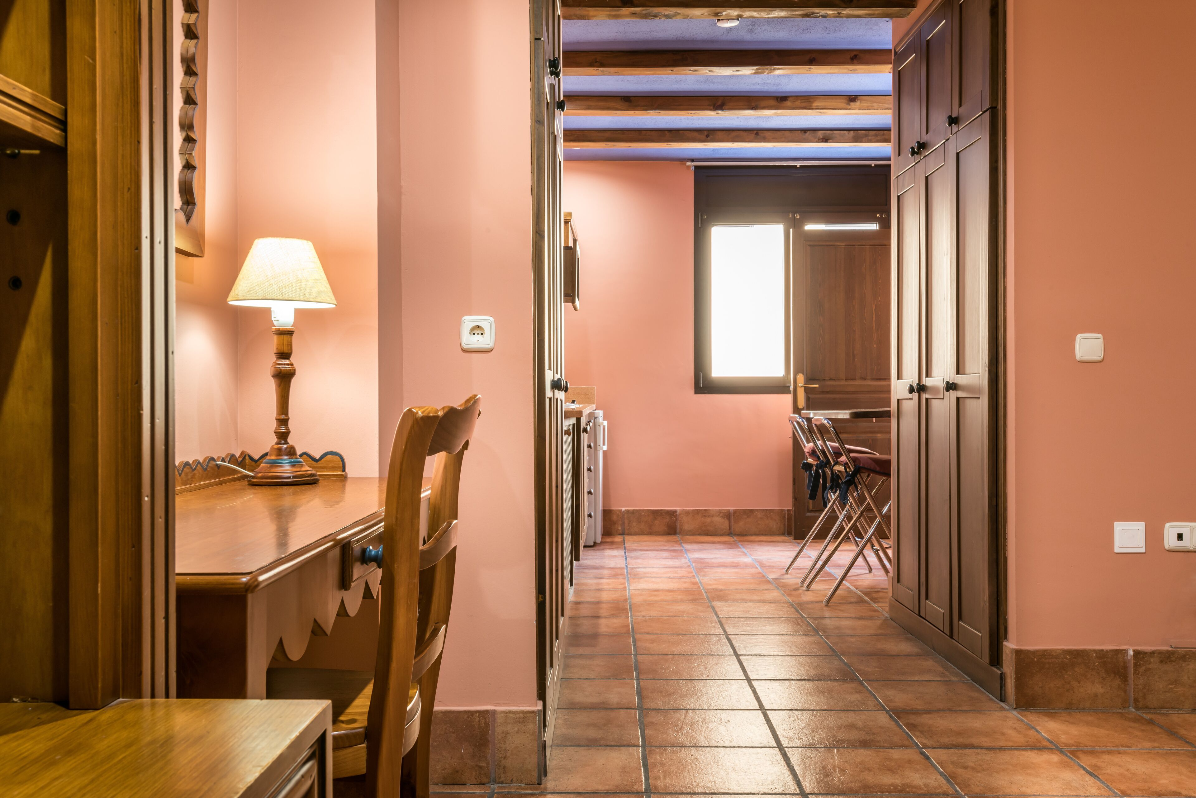 A narrow hallway with a wooden table and chairs, a lamp, and a window at the end.