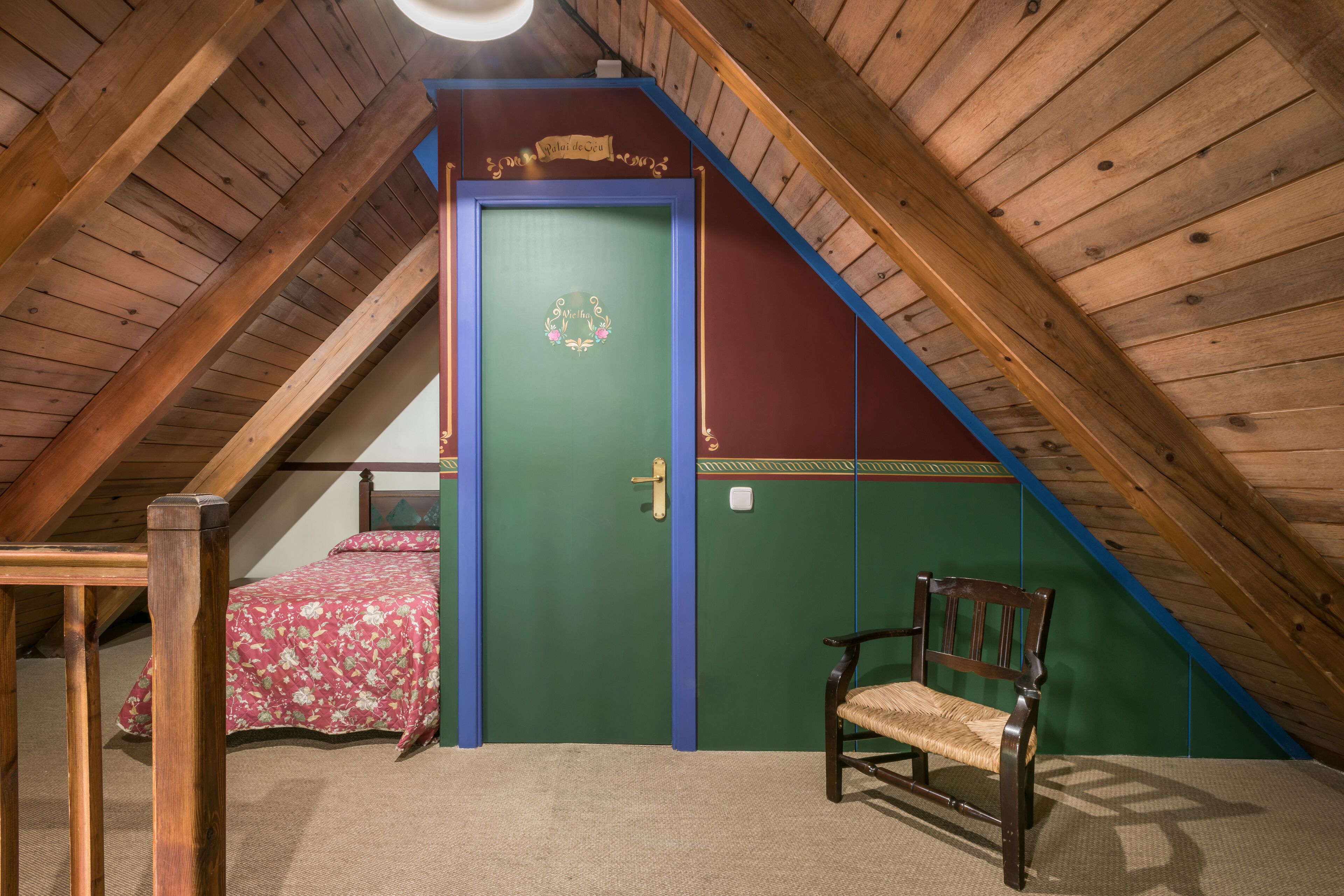 Une pièce avec un plafond en pente, une porte verte ornée d’une guirlande décorative, un lit recouvert d’une couvre-lit à motifs floraux et un banc en bois.