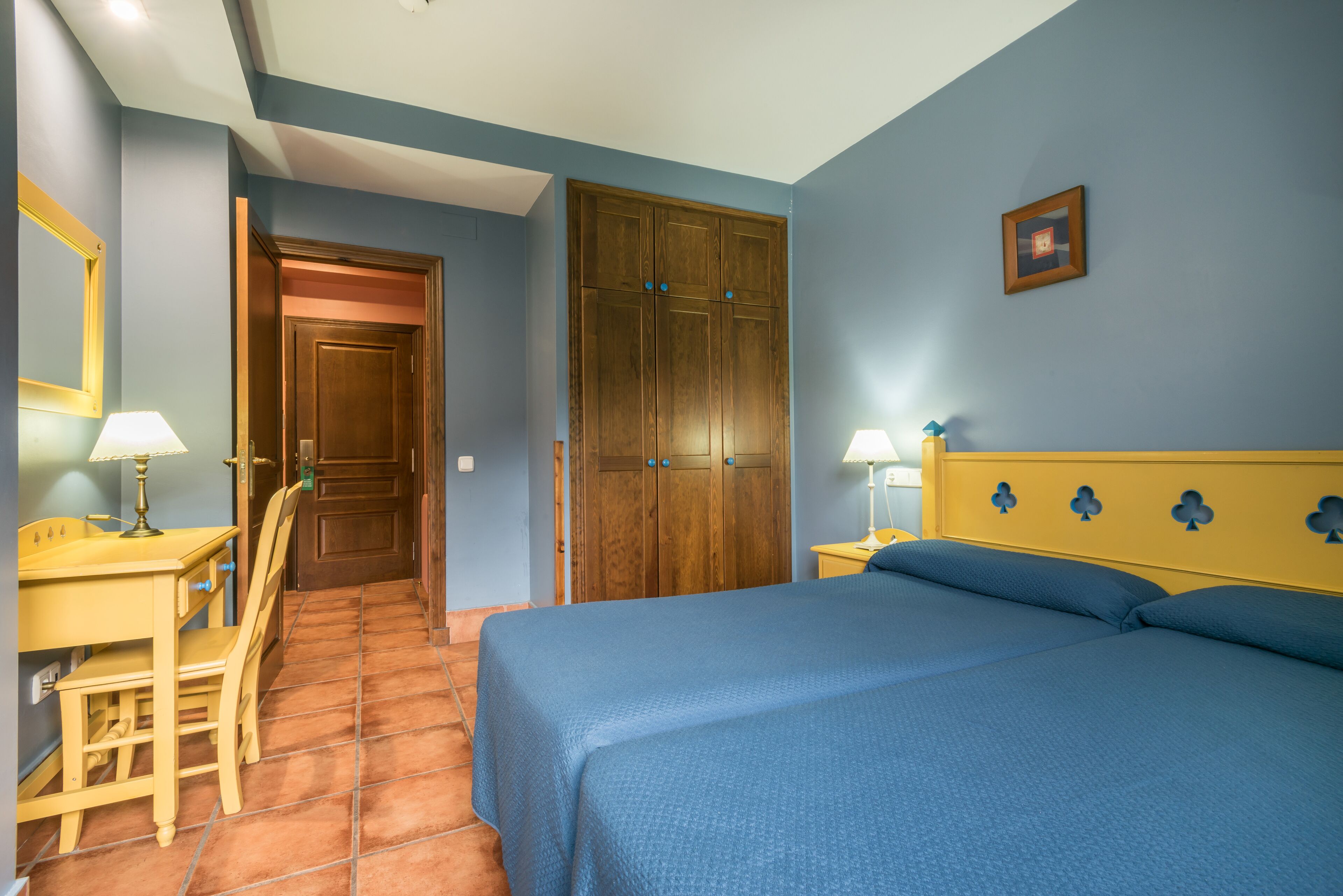 A bedroom with a bed, a desk, a chair, a wardrobe, and a bedside table with lamps.
