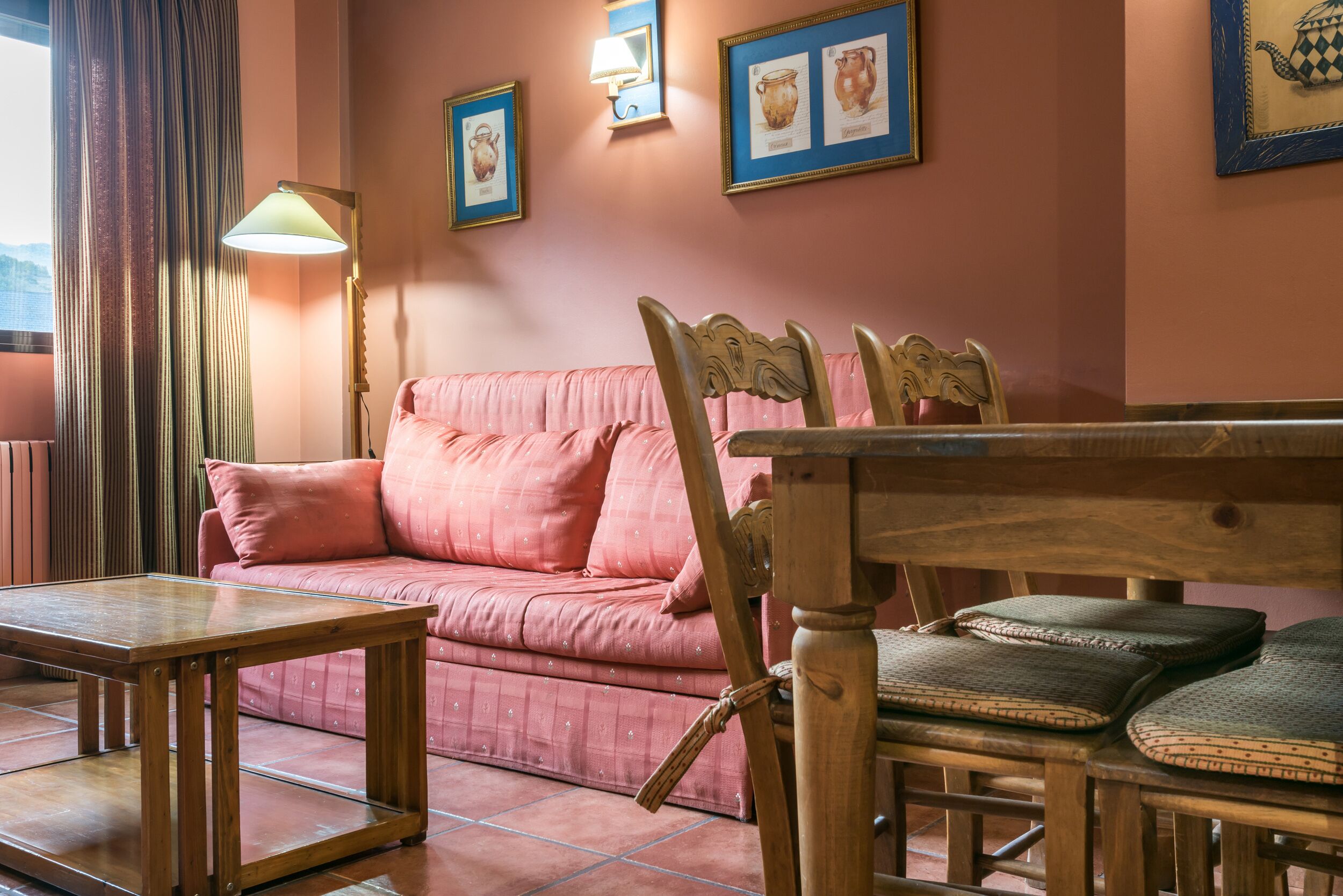 Un salón acogedor con un sofá rosa, sillas de madera, una mesa de centro de vidrio y cuadros en la pared.
