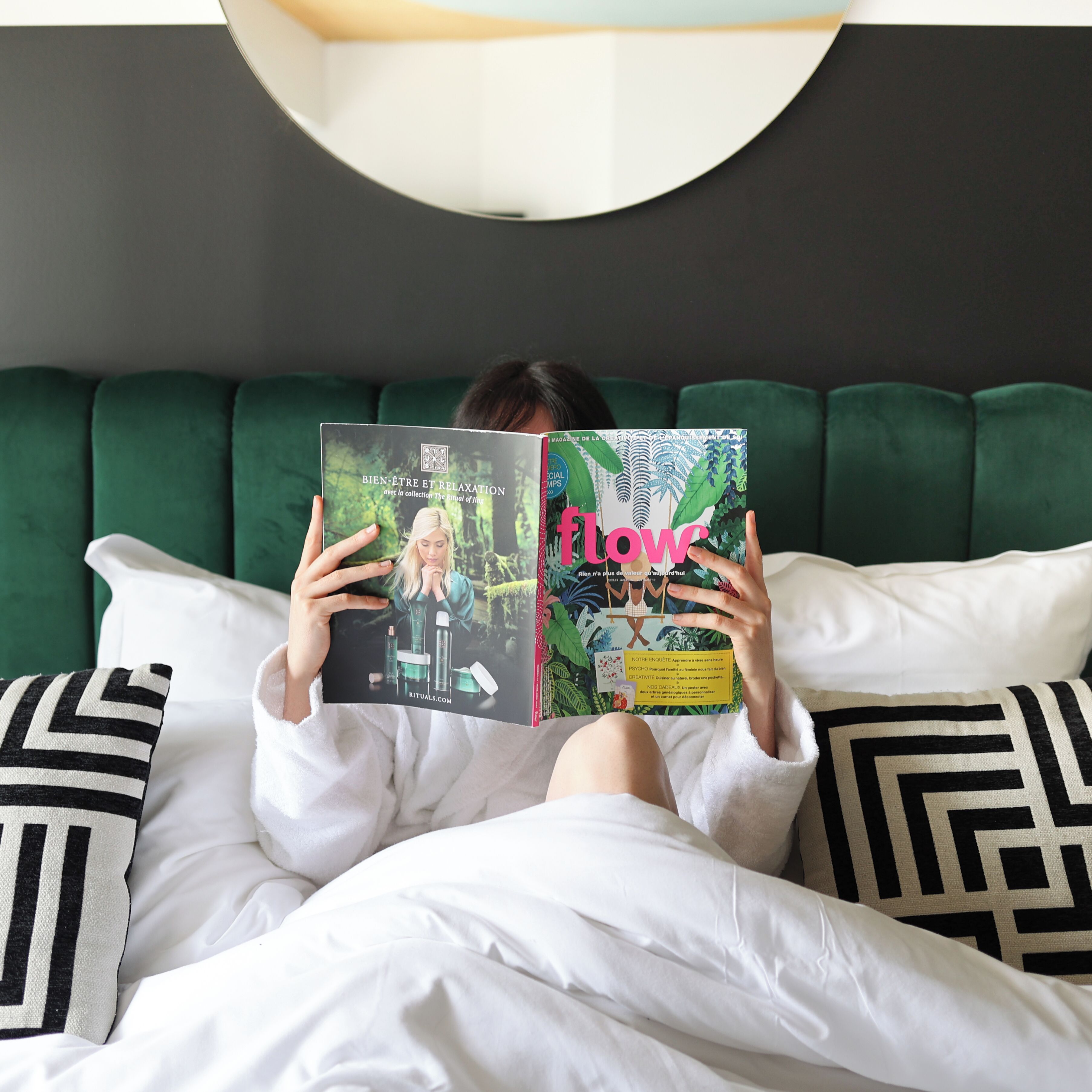 A person lying on a bed with a magazine, pillows, and a green headboard.