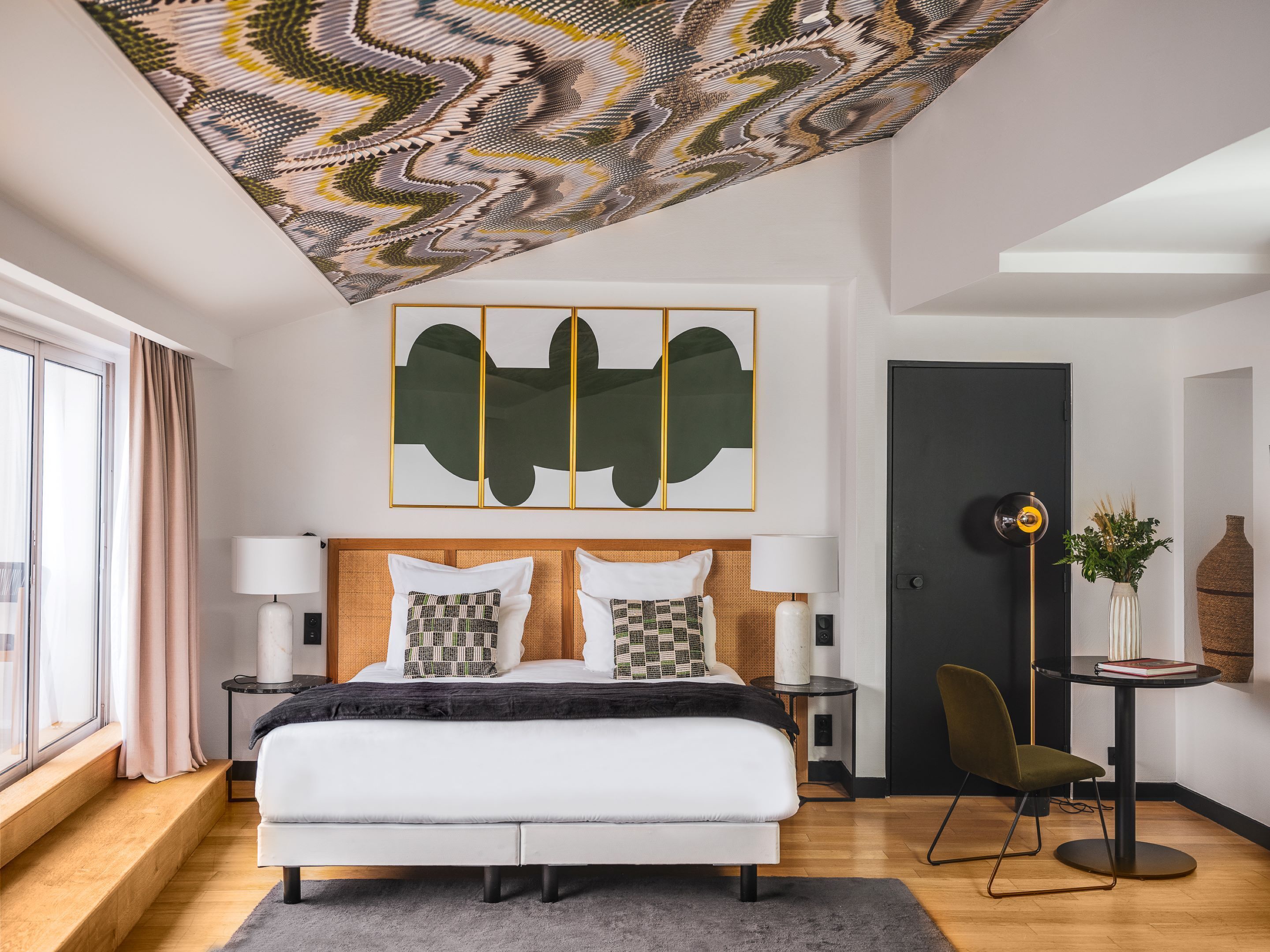 Quarto moderno com cama grande, obra de arte no teto, porta escura e pequena mesa com planta.