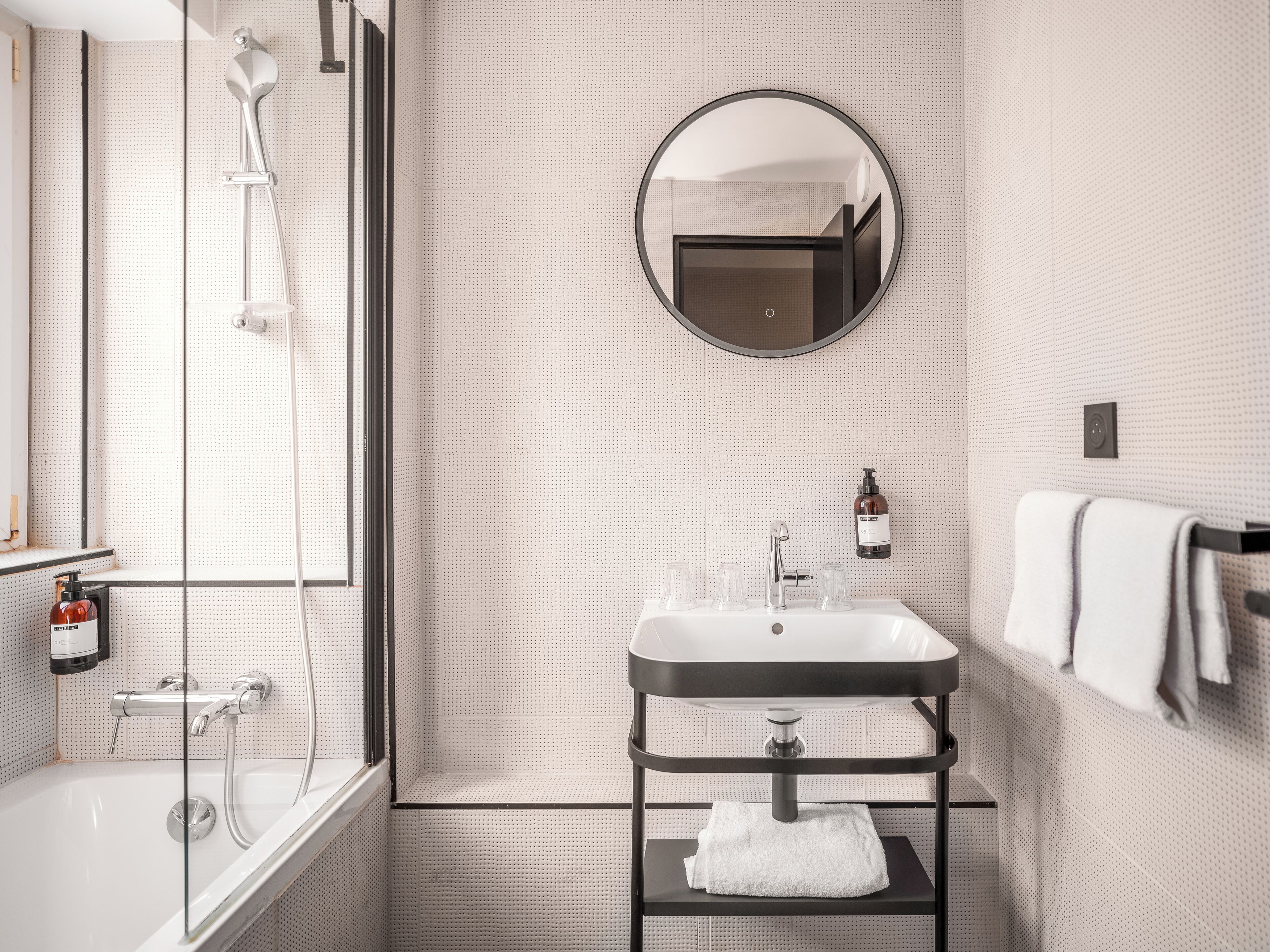 Un baño moderno con ducha de vidrio, lavamanos blanco sobre encimera negra, espejo redondo y barra para toallas.