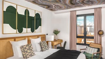 A hotel room with a large bed, a desk, and a chair. There is a view of the city through the window.