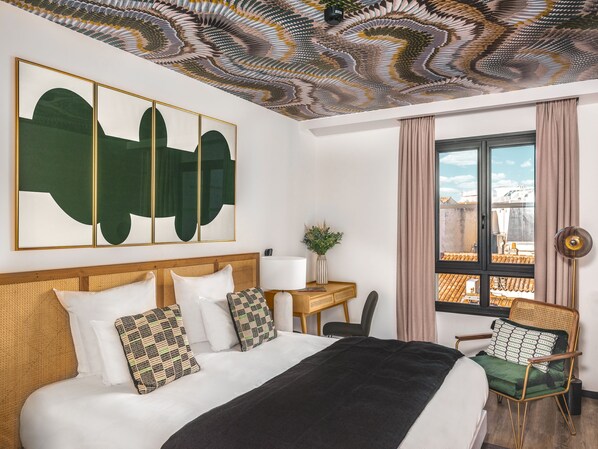 A hotel room with a large bed, a desk, and a chair. There is a view of the city through the window.