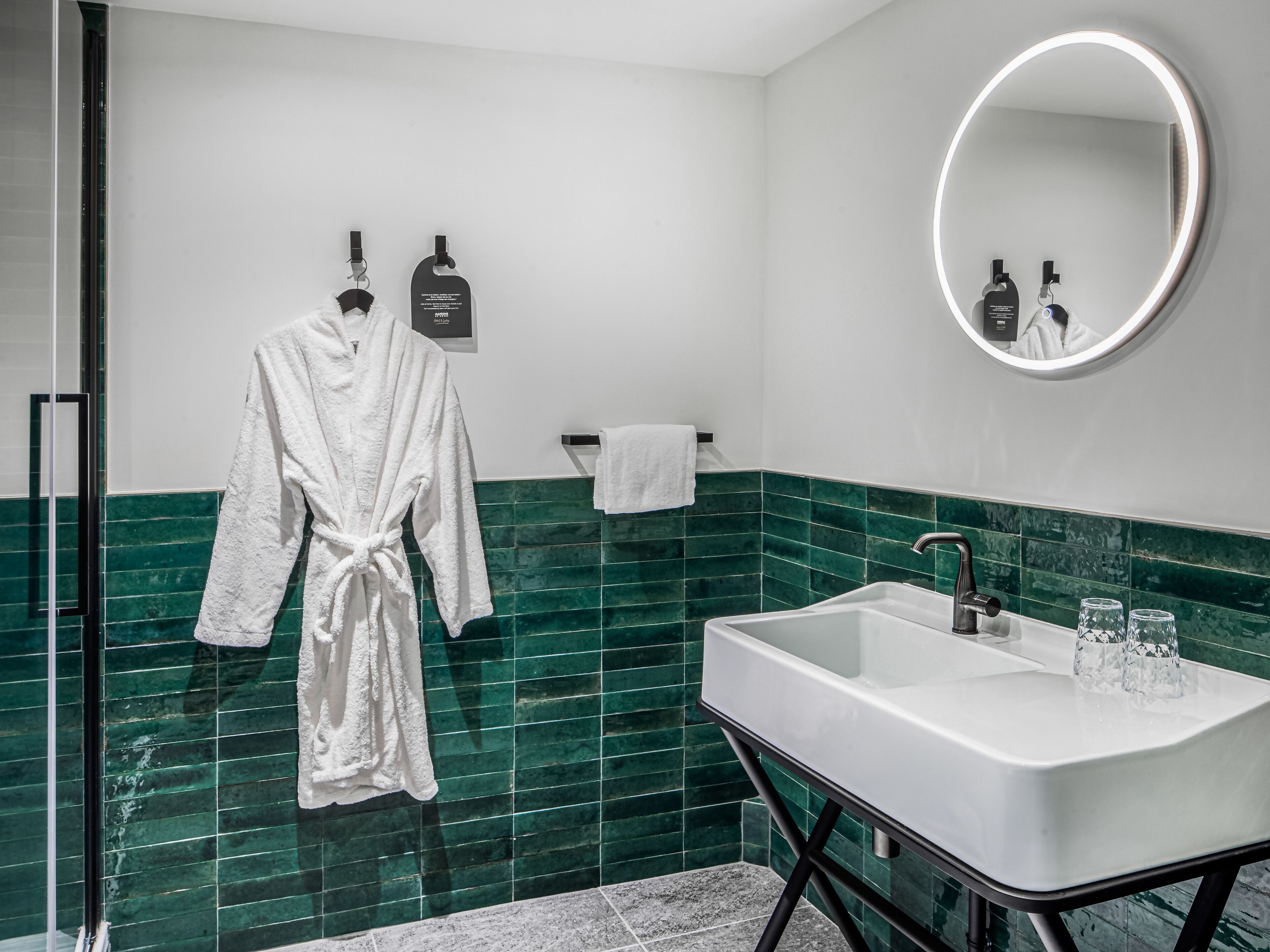 Un baño moderno con un lavabo blanco, baldosas verdes, un espejo redondo y una bata blanca colgada en ganchos.