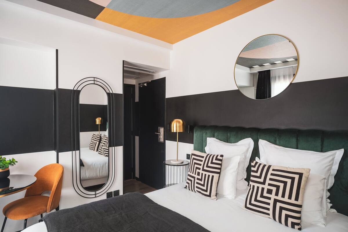 Una habitación de hotel moderna con una cama grande, un espejo redondo, un cabecero tapizado en verde y una mesita de noche con una lámpara.