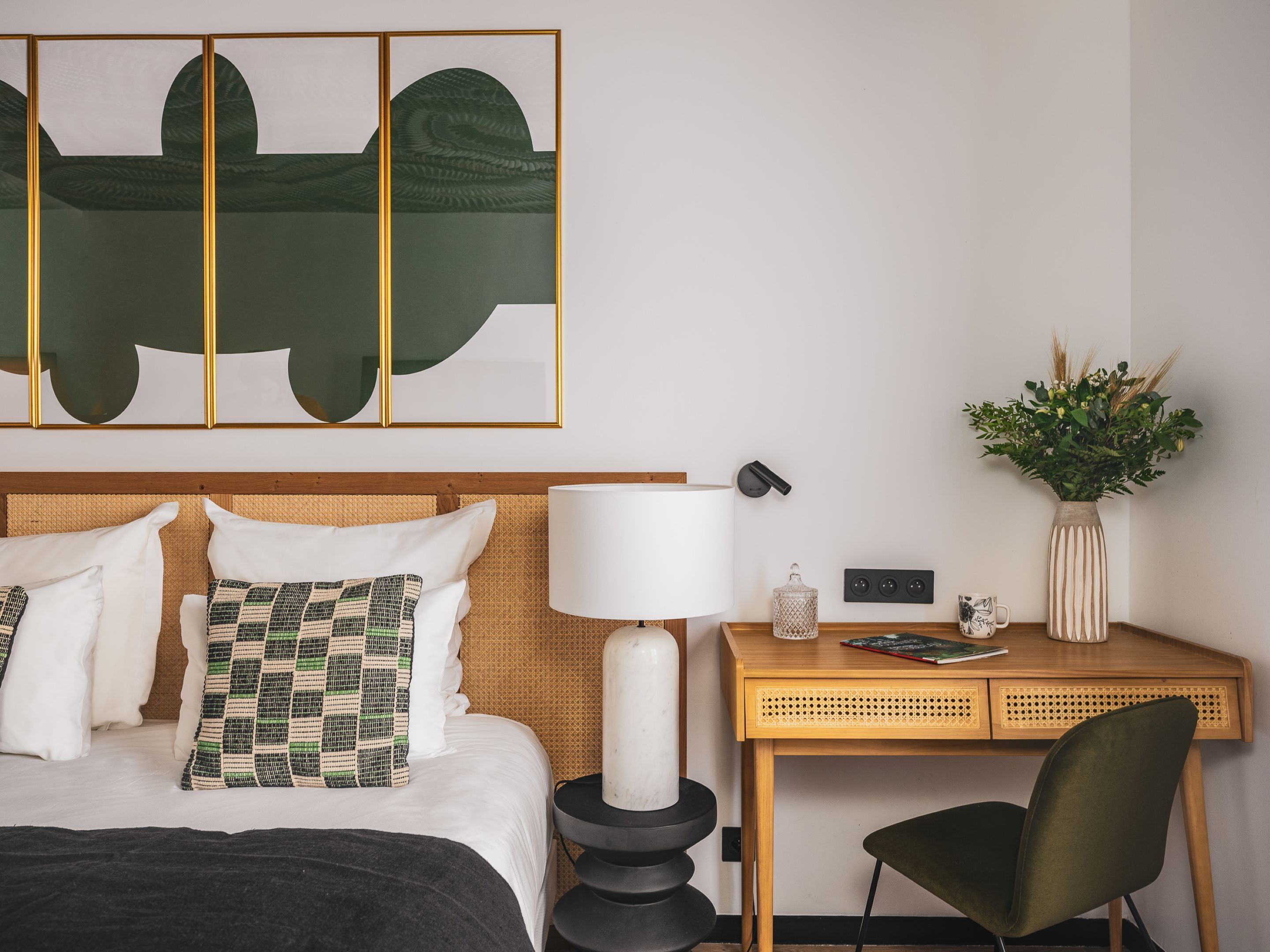 Ein Hotelzimmer mit Bett, Schreibtisch, Stuhl, Lampe, einer Blumenvase und einem Wandbild.