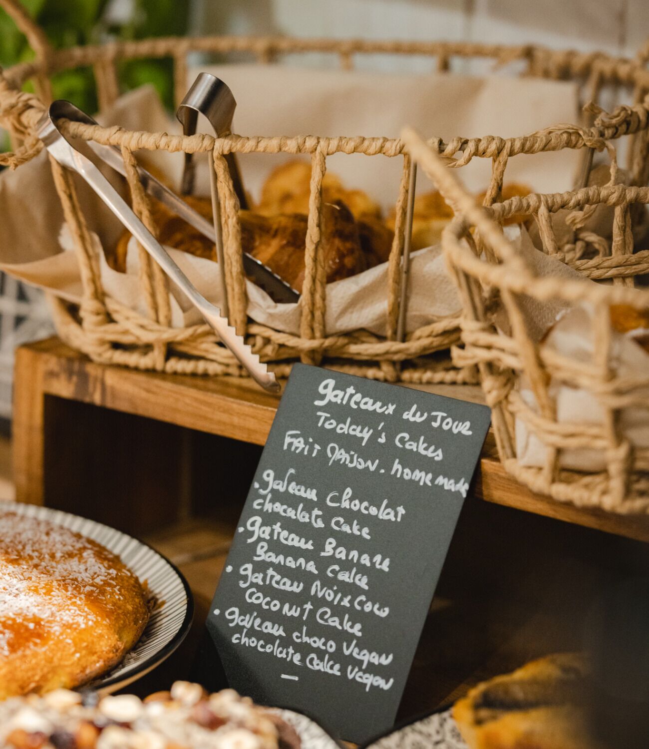 A menu board listing various cakes and desserts, a basket with baked goods, and a plate of cake with powdered sugar.
