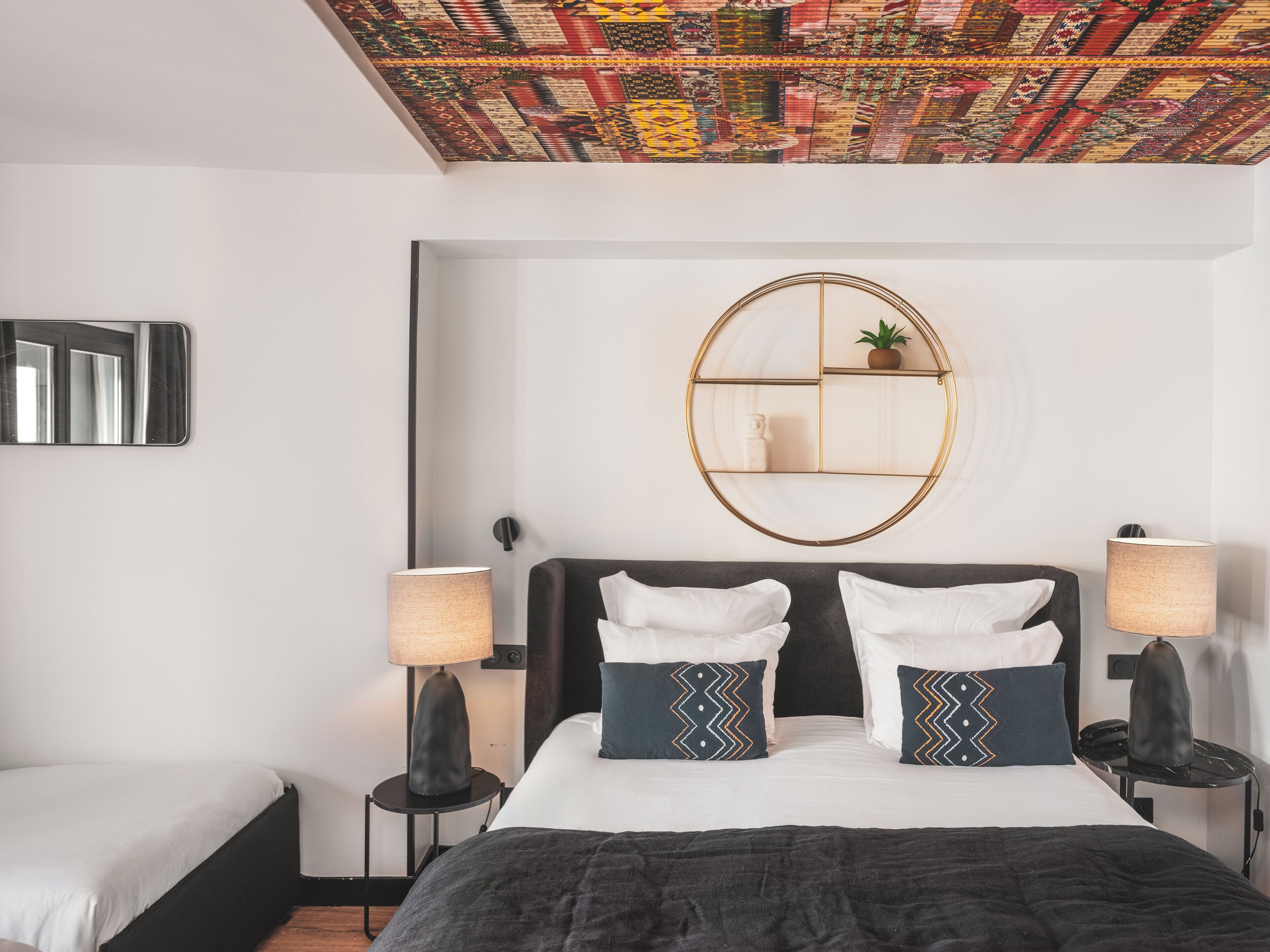 Una camera da letto moderna con un letto grande, due comodini con lampade, uno specchio rotondo e un soffitto con un motivo colorato.