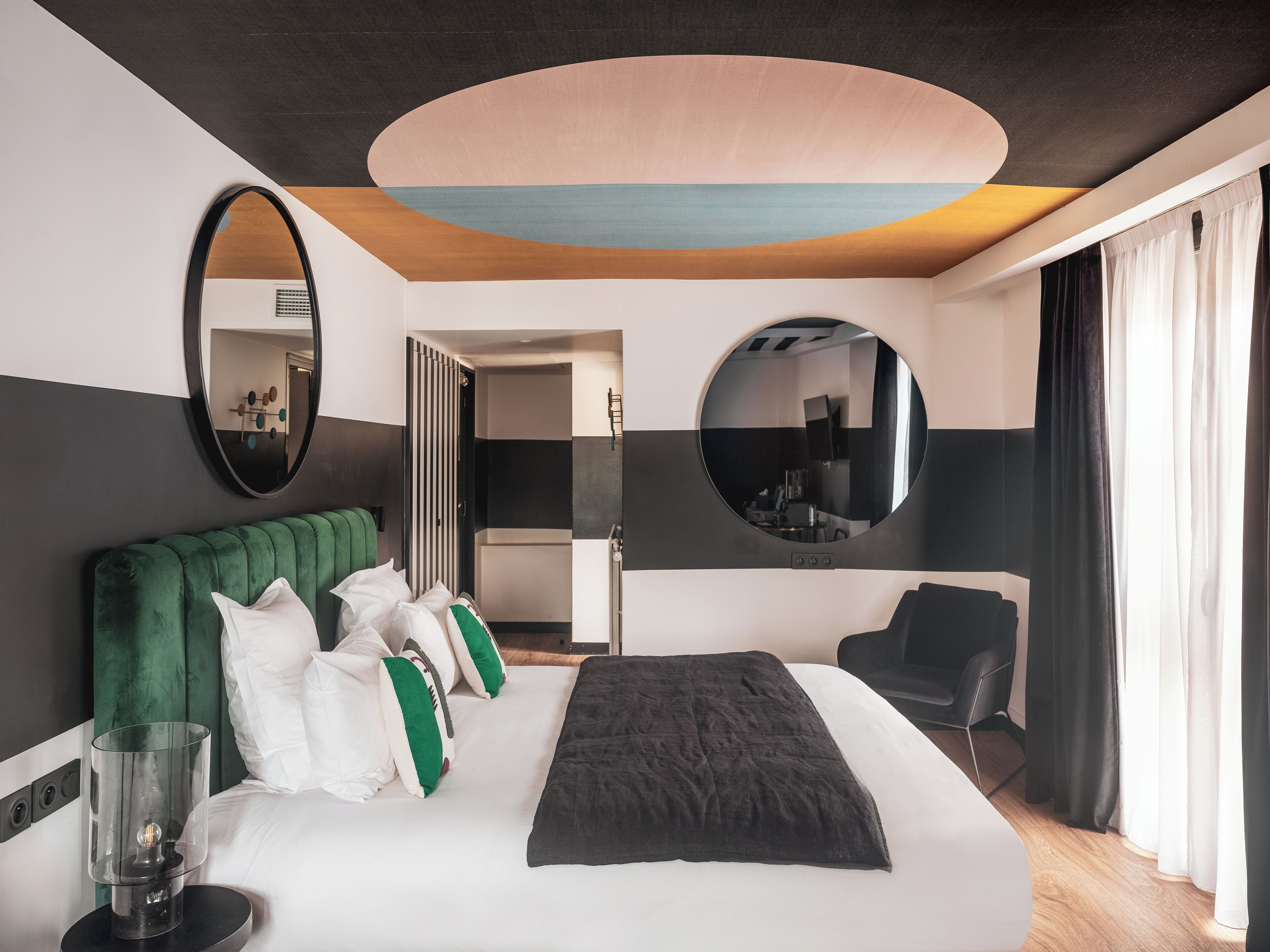 A modern hotel room with a large bed, a green upholstered headboard, a round mirror, and a view of the room's interior through a circular mirror.