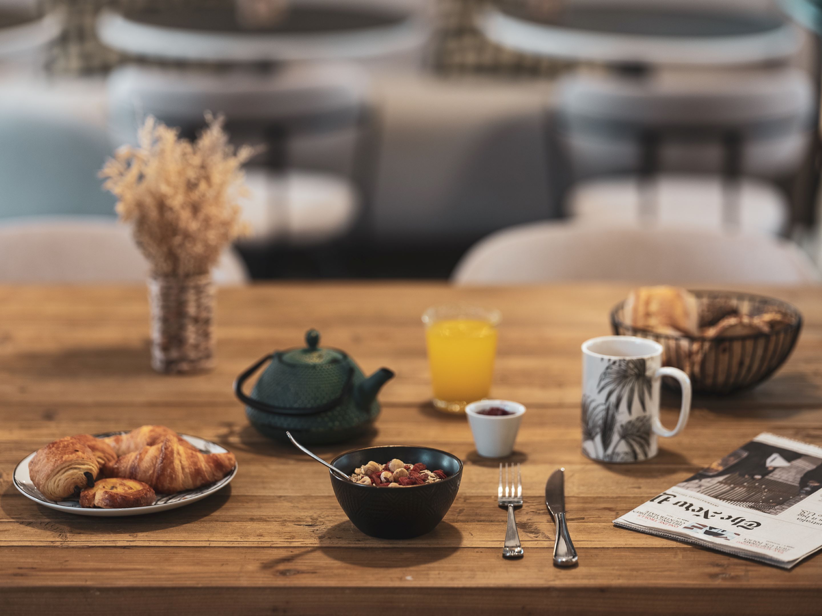 A breakfast table with croissants, a bowl of cereal, a teapot, a glass of orange juice, a coffee mug, and a newspaper.