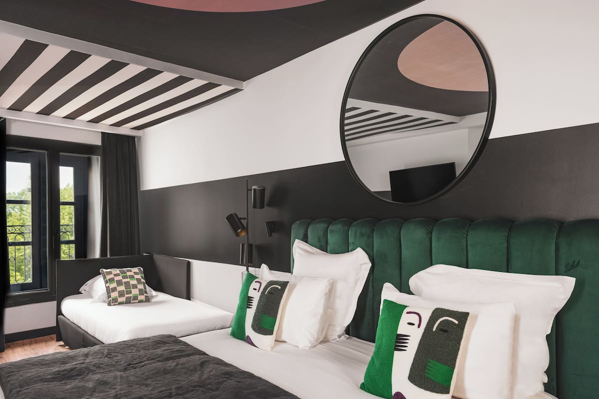 Una habitación de hotel moderna con una cama grande, dos mesitas de noche, un cabecero tapizado en verde y un espejo grande, además de una ventana con vistas a zonas verdes.