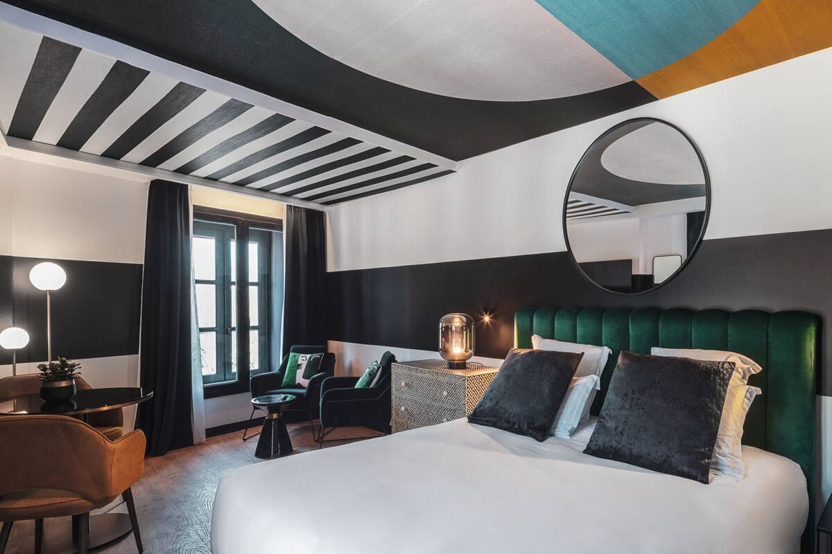 Una habitación de hotel moderna con una cama grande, un espejo redondo, un cabecero tapizado de verde y una pequeña mesa con una lámpara.