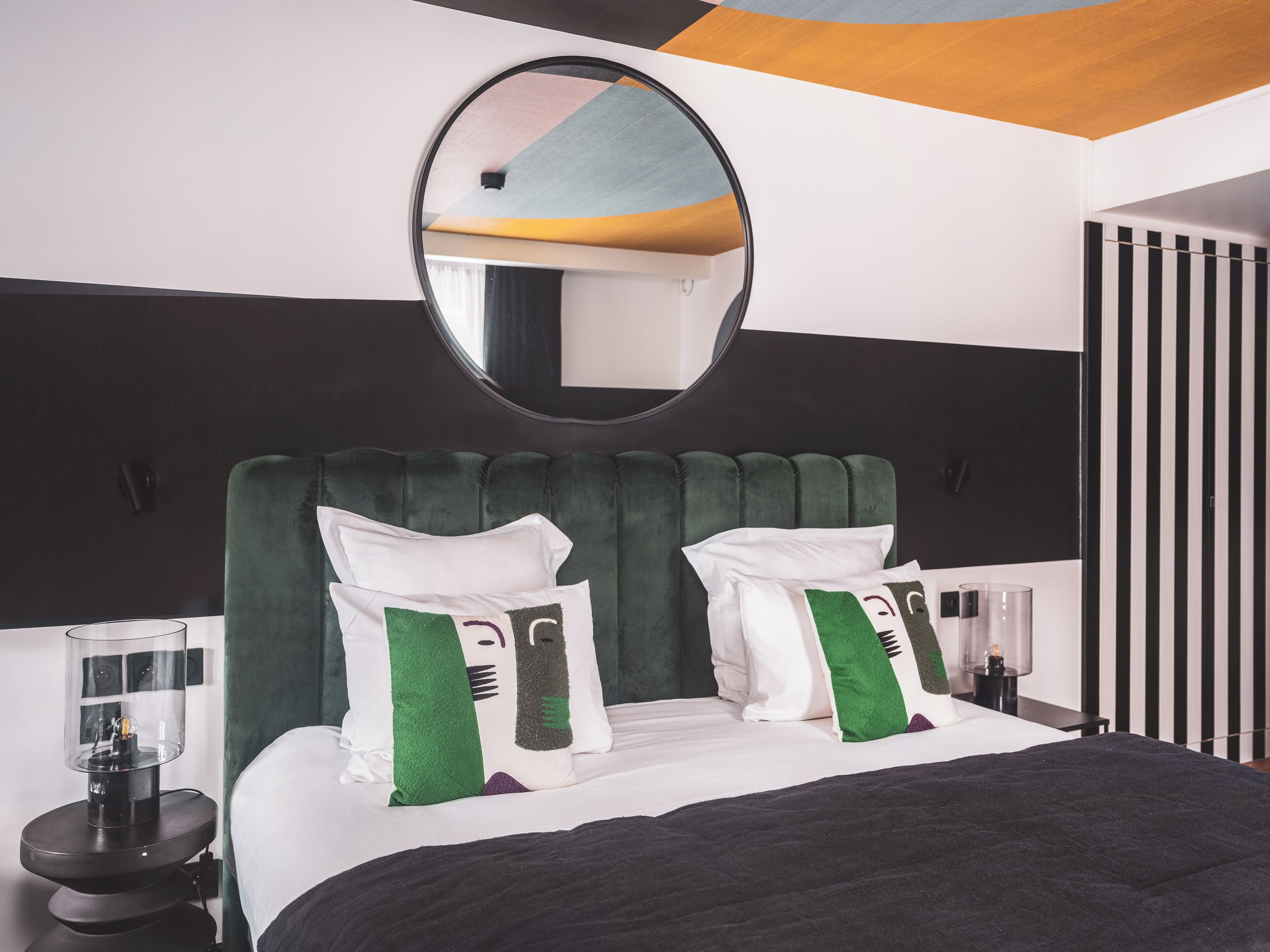 Una habitación de hotel moderna con una cama grande, cabecera tapizada en verde y un espejo redondo encima.
