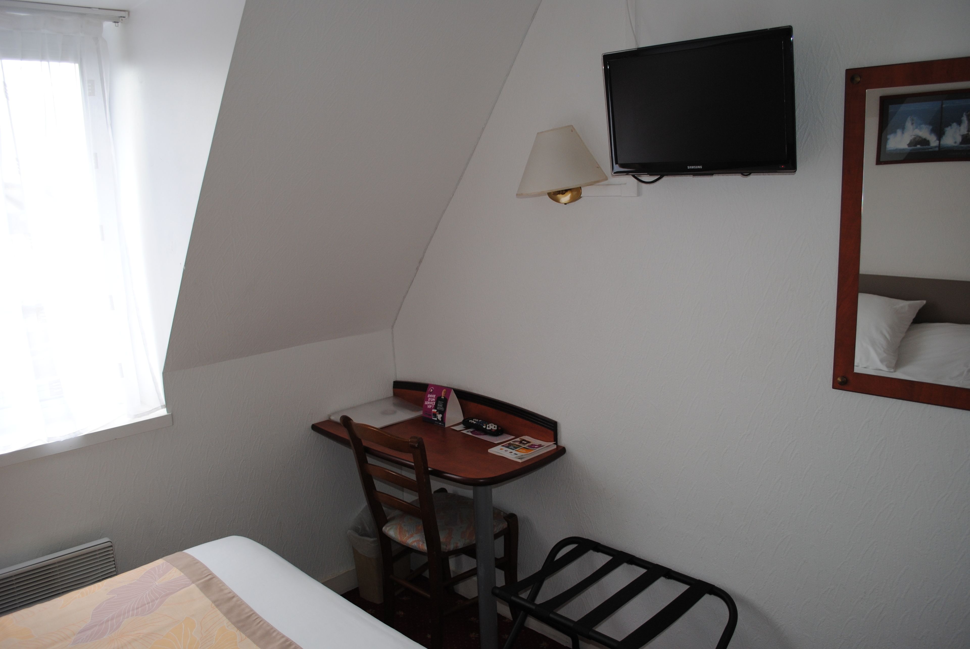 A small, well-lit room with a bed, a desk with a chair, a television, and a window with curtains.