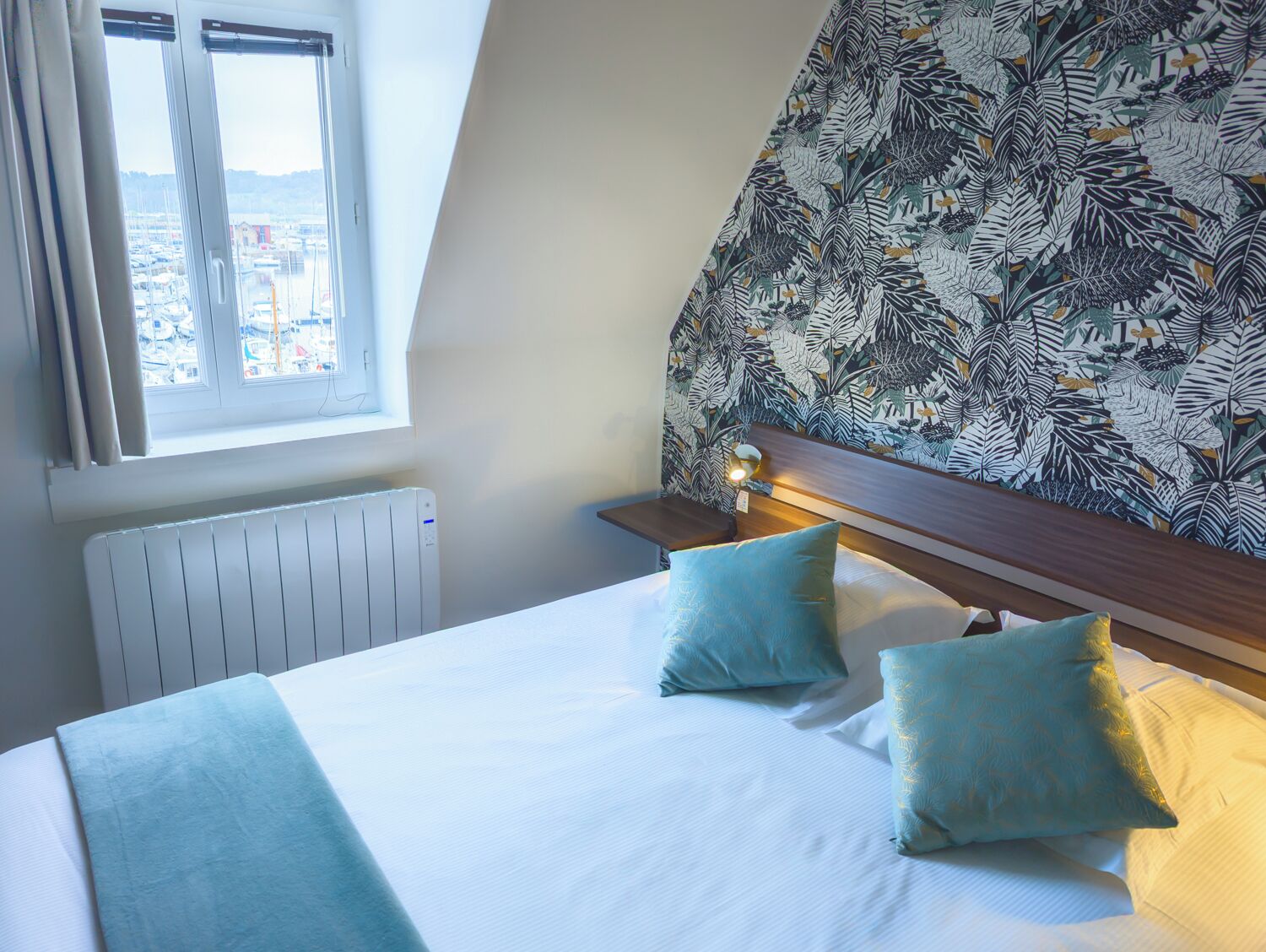 A hotel room with a bed, two pillows, a nightstand with a lamp, a radiator, and a window with a view of buildings.