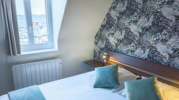 A hotel room with a bed, two pillows, a nightstand with a lamp, a radiator, and a window with a view of buildings.