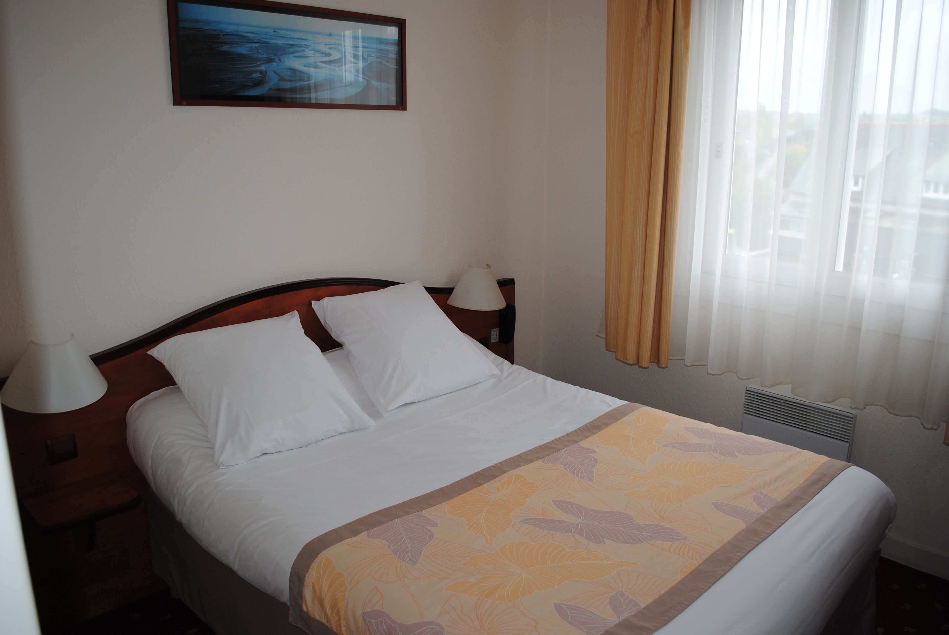 A hotel room with a bed, a nightstand, a lamp, a painting on the wall, and a window with curtains.