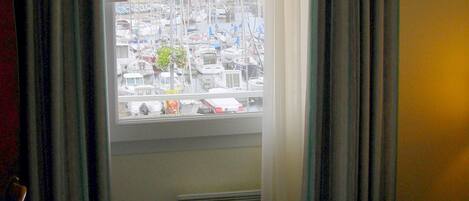 Habitación de hotel con cama, ventana con cortinas y vista a un puerto deportivo con barcos.