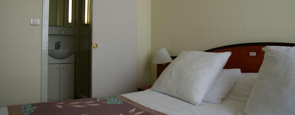 Una habitación de hotel con cama, mesita de noche con lámpara, tocador y baño con lavabo y espejo.