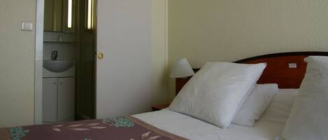Una habitación de hotel con cama, mesita de noche con lámpara, tocador y baño con lavabo y espejo.