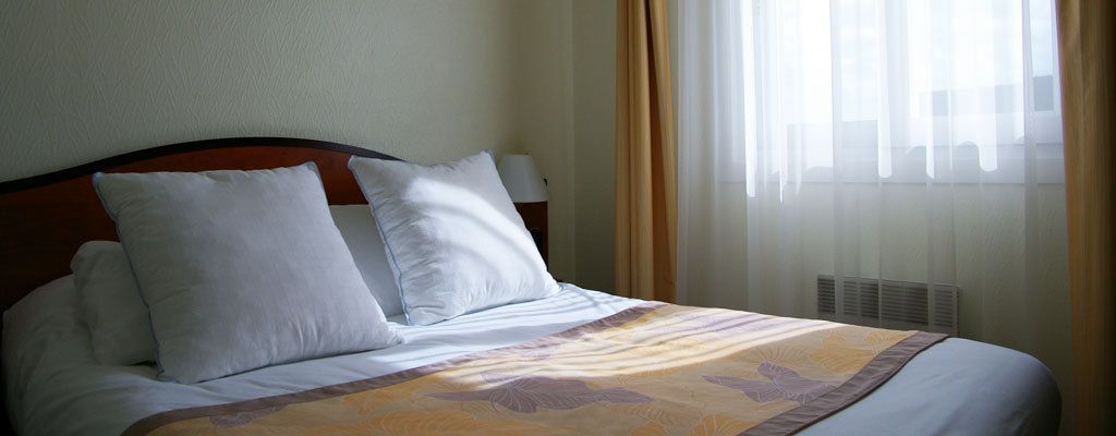A hotel room with a bed, white pillows, a headboard, a bedside table with a lamp, and a window with curtains.