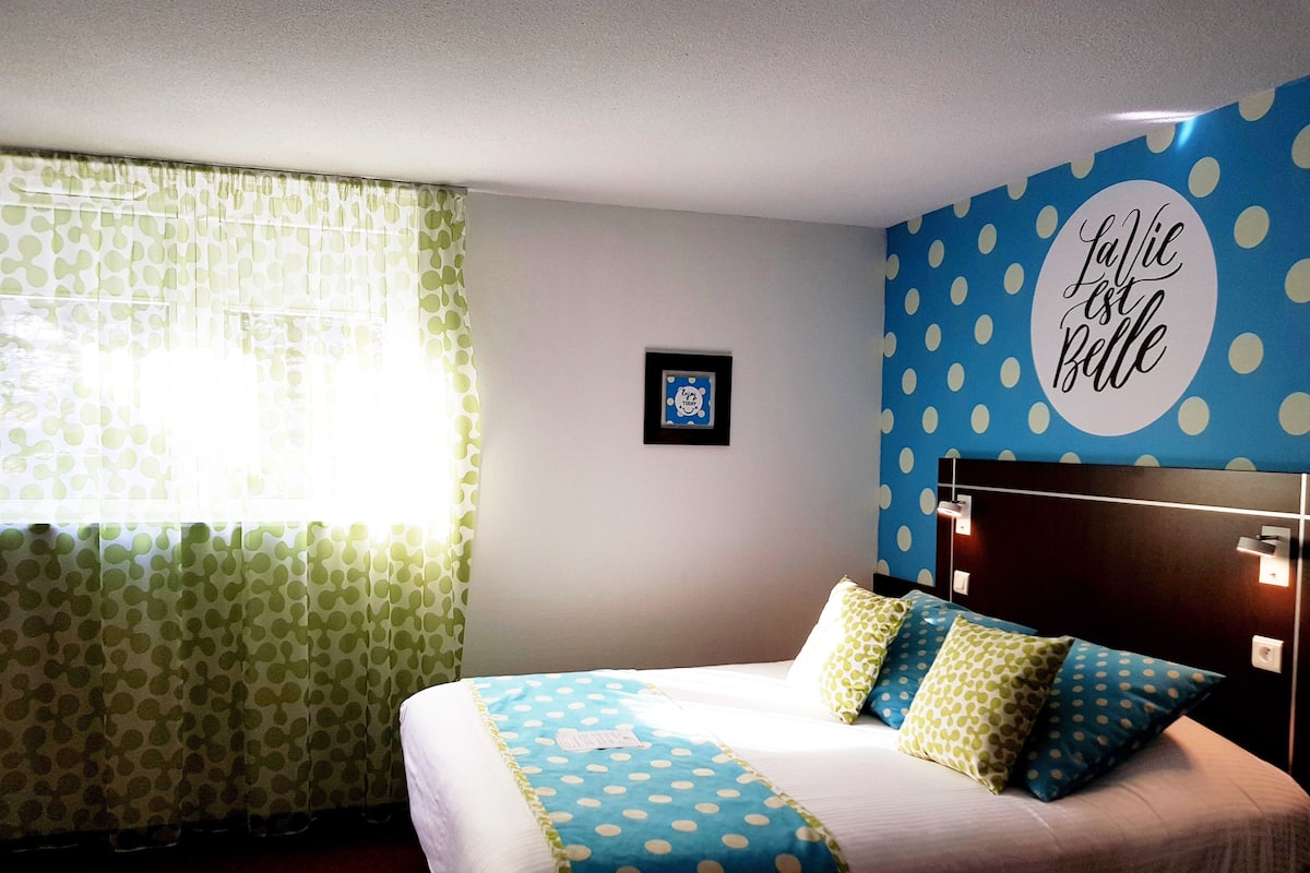 A hotel room with a bed, a window with green curtains, a wall with a blue polka dot pattern, and a framed picture on the wall.